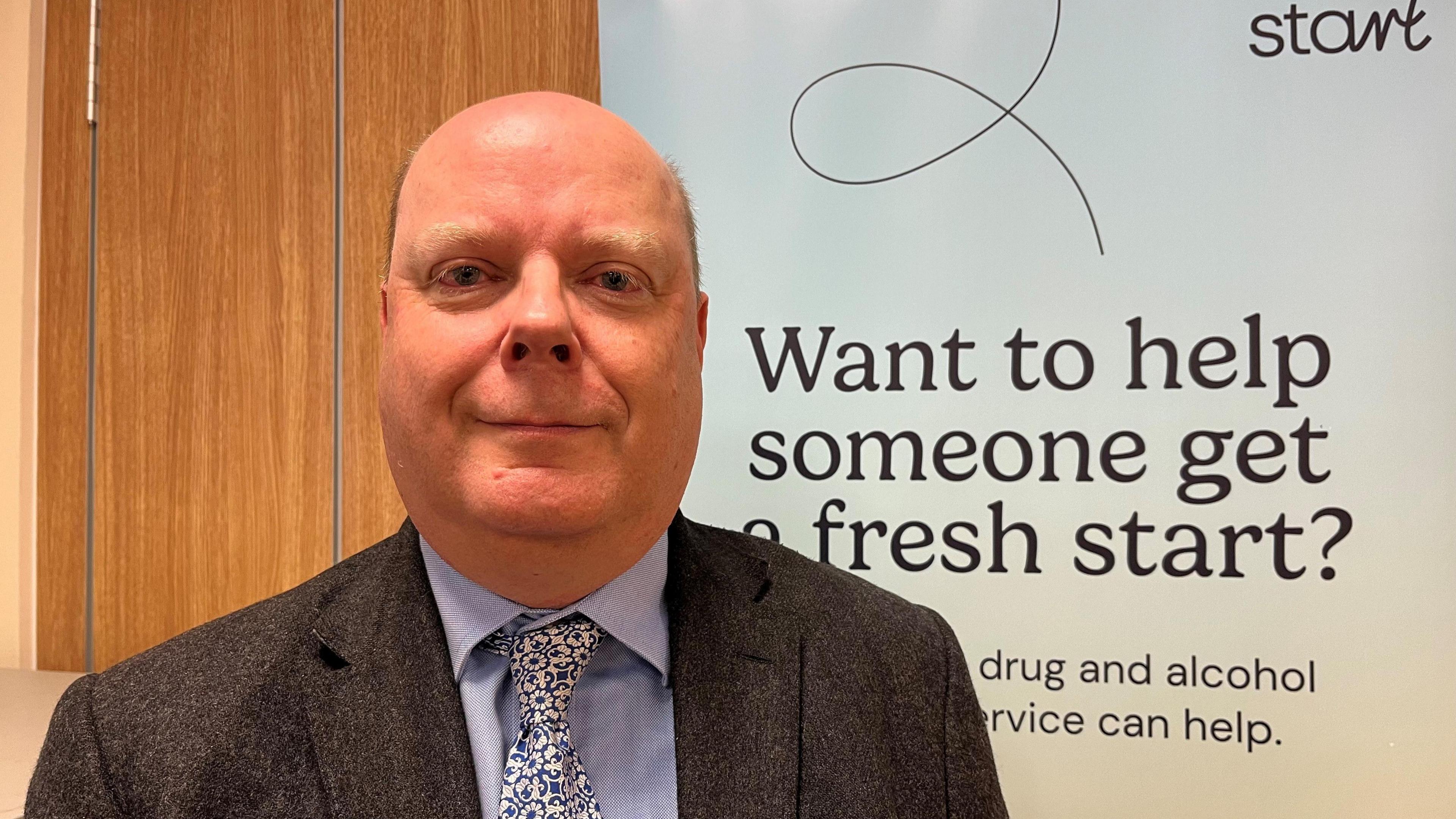 Craig Blundred, Director of Public Health, Hartlepool Borough Council, standing in front of a drug advice poster, slightly smiling, wearing a suit and tie.