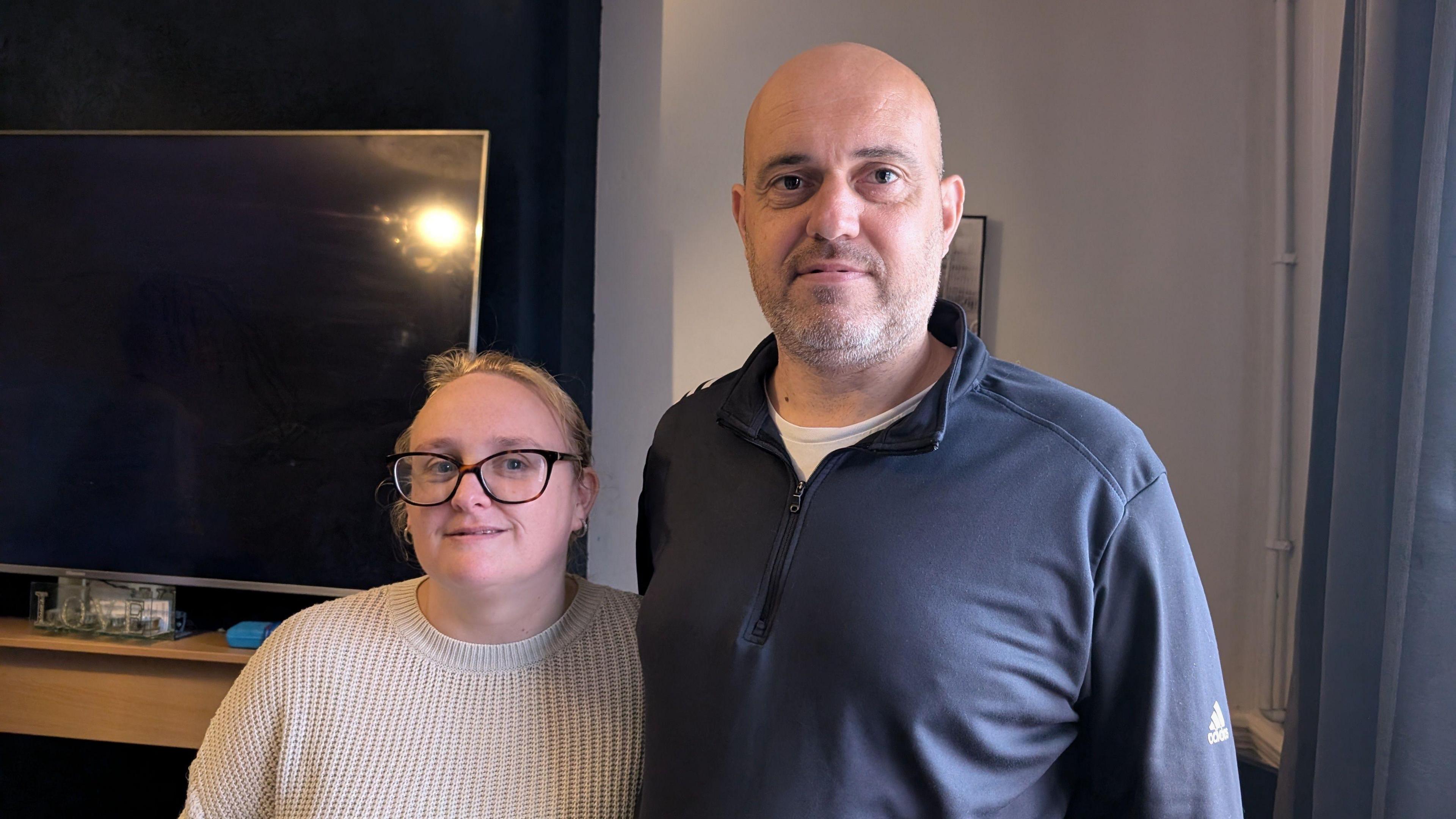 Charlotte and Carl Cooper. Charlotte is wearing a beige knitted jumper, Carl is wearing a grey jumper over a white t-shirt. There is a large television on the wall behind them and blue curtains to the right of them.