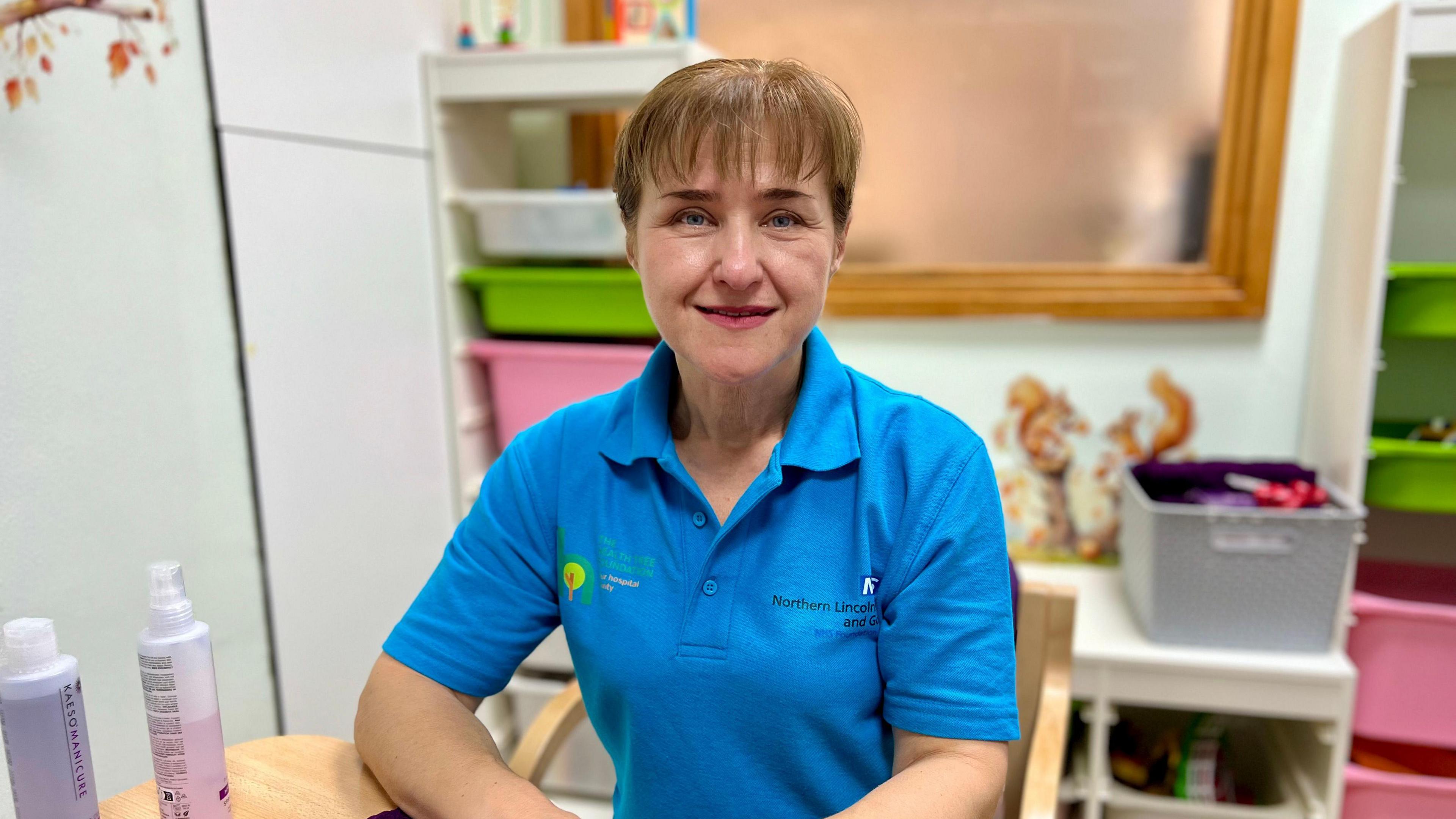 A person in a blue polo shirt with the Northern Lincolnshire and Goole NHS Trust logo sits at a table. There are coloured boxes in the background.