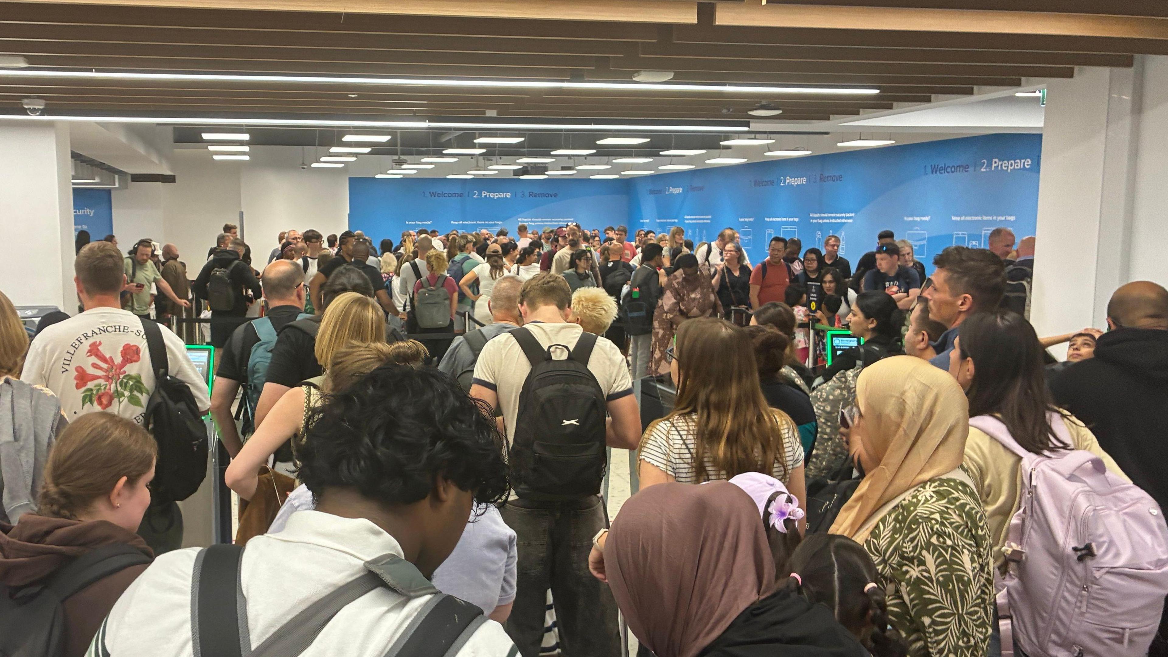 Crowds of people queue at Birmingham airport. You can see the back of people's heads as they stand still. 