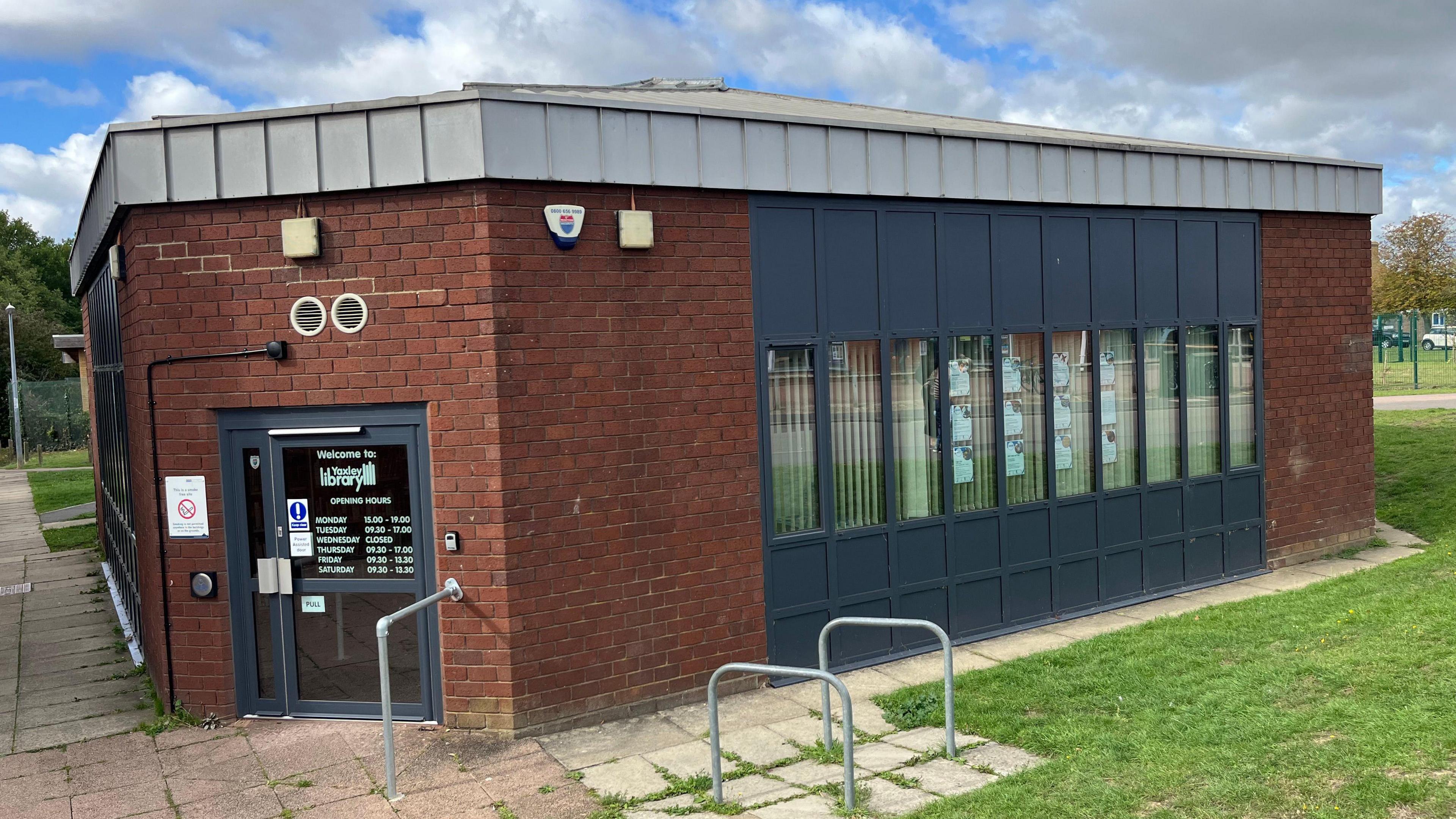 The exterior of the small building which stands on its own surrounded by grass. Its one storey and made of red brick with some black cladding around the windows.