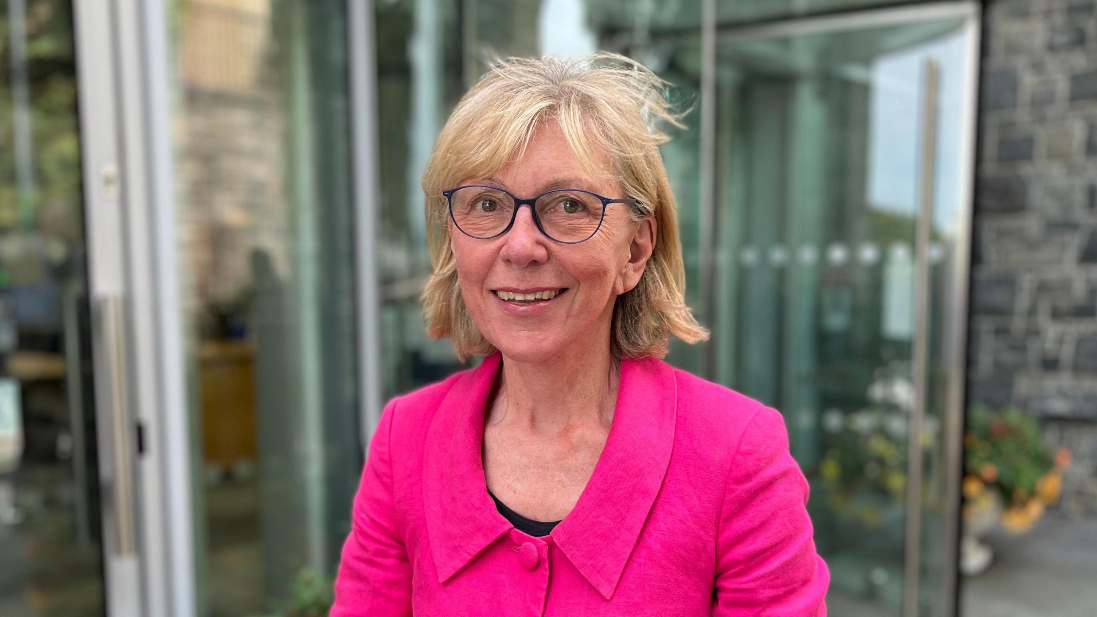 Deputy Yvonne Burford - A woman with mid-length blonde hair and blue rimmed spectacles. She's wearing a bright pink jacket on top of a black blouse.