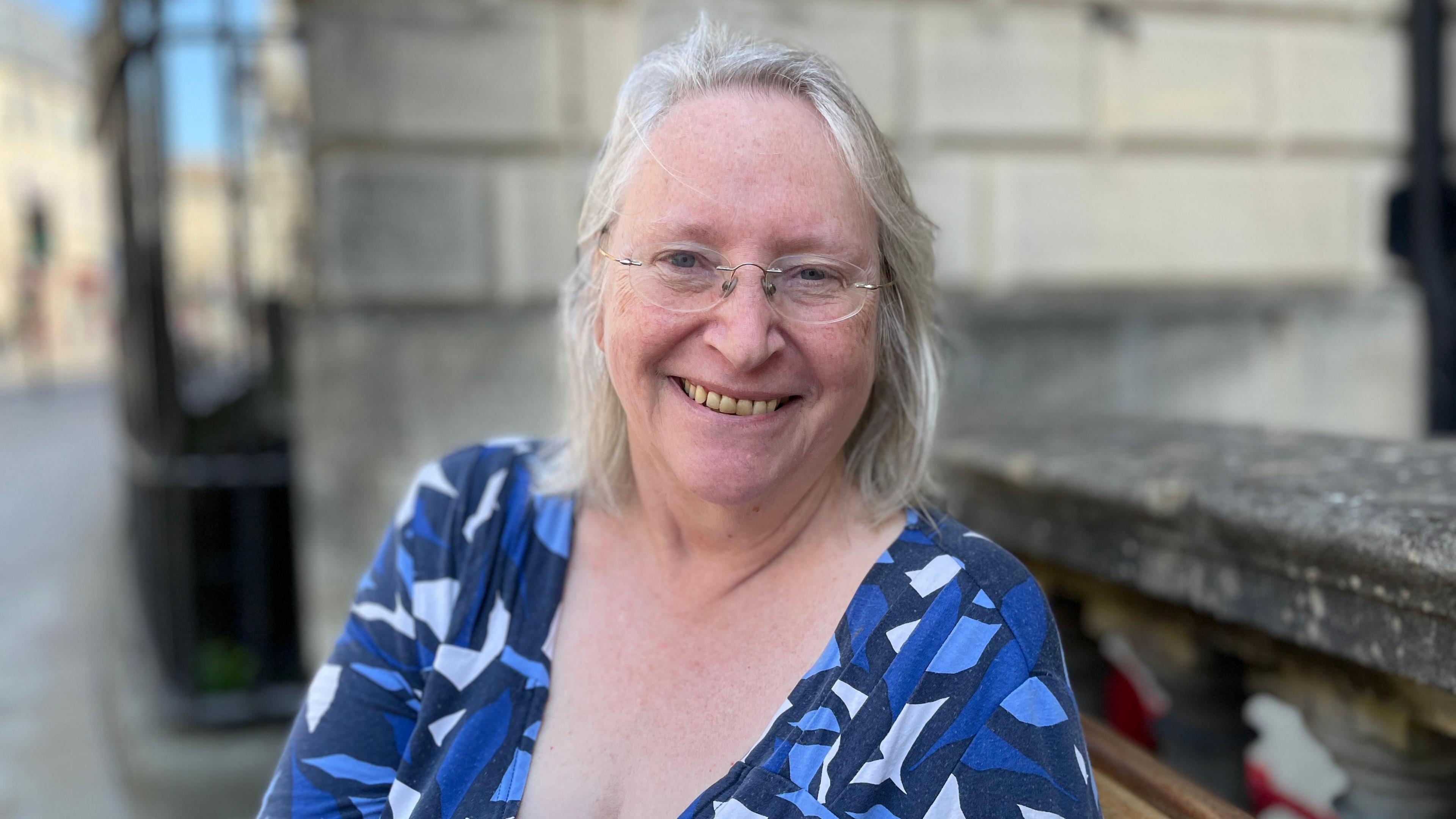 Manda Rigby sits smiling into the camera. She has grey and white shoulder-length hair, and is wearing a patterned blue top. She wears rounded glasses.