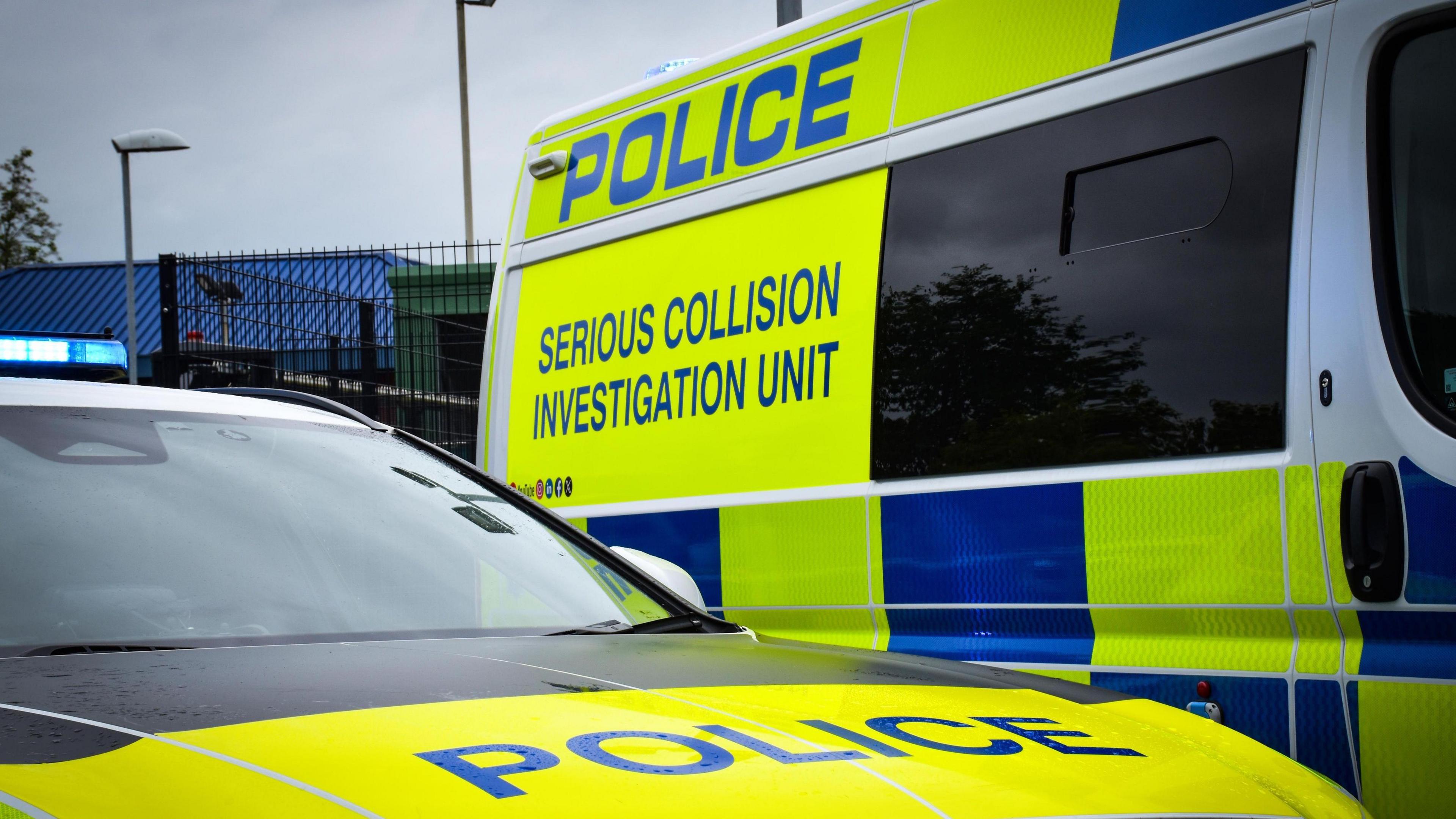 A police car to the front of the camera, and a police van parked behind it, with the wording 'serious collision investigation unit' on the side of the vehicle