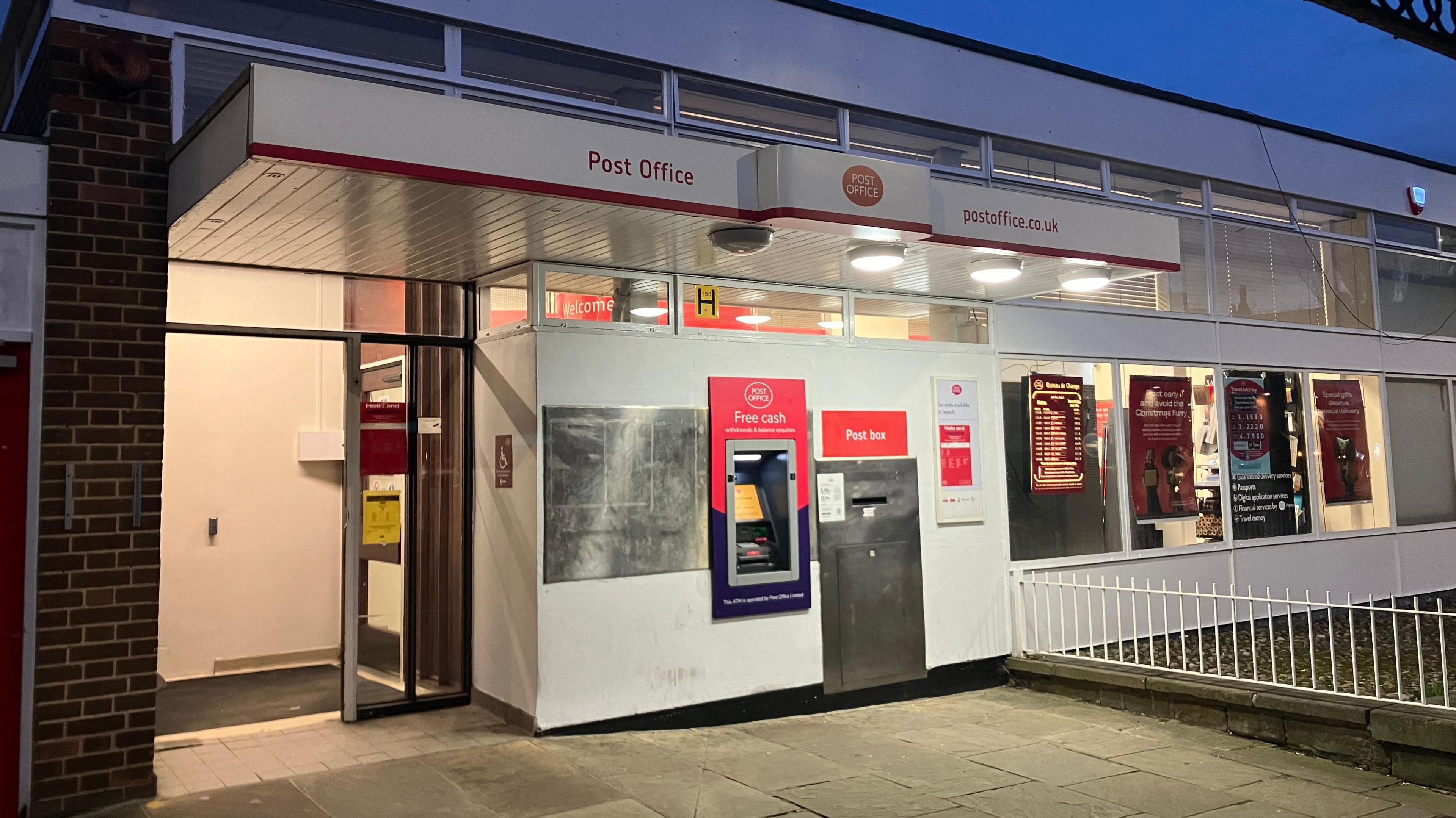 The entrance to the post office which features an ATM and a post box on the right. Several posters hang in the window as well as a currency conversion board.