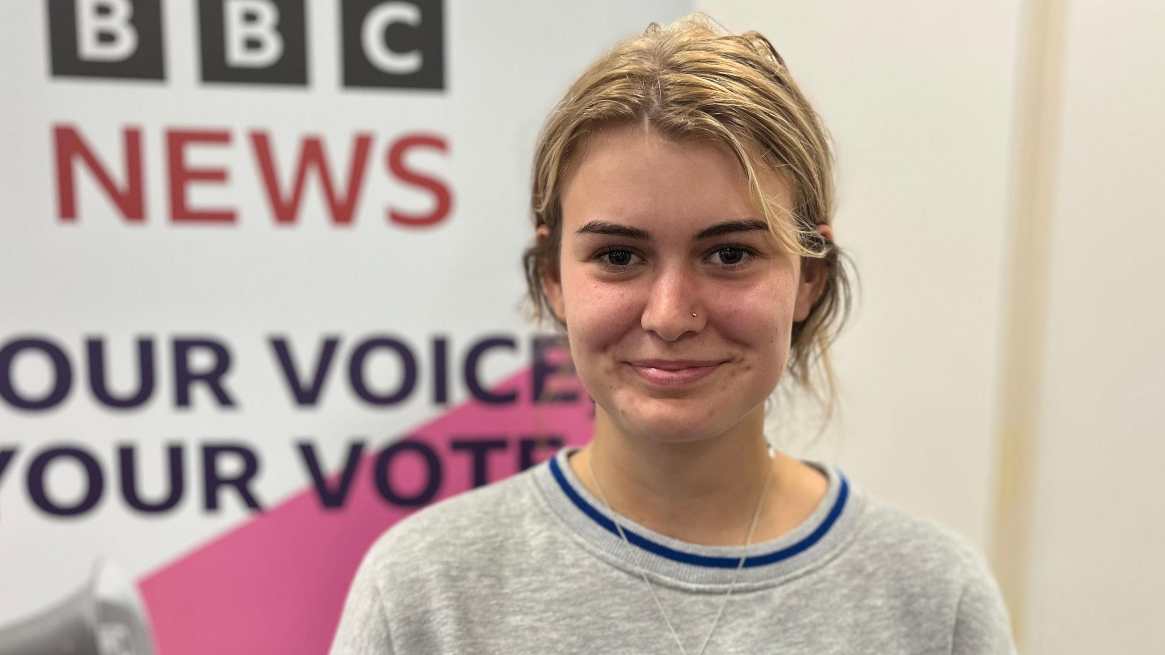 A teenage girl with blonde hair and brown eyebrows. She's wearing a grey jumper and a gold chain necklace.