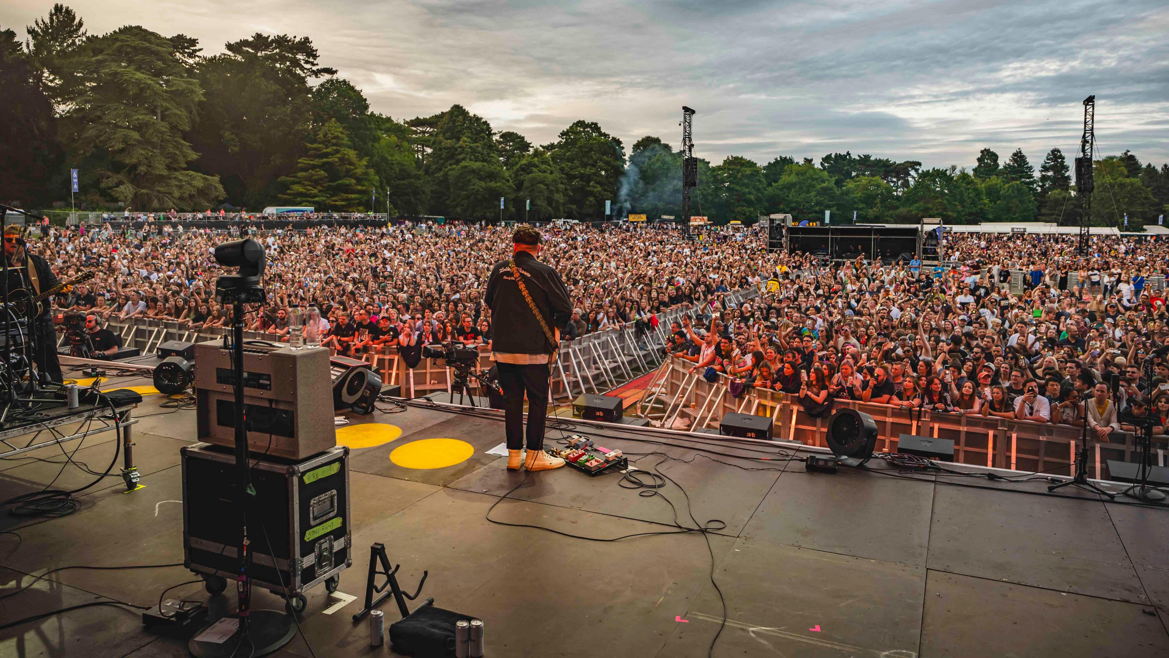 A concert in Bedford Park, showing someone on stage, playing a guitar and a large crowd in front . There are trees in the distance. Sound equipment is on the stage and leads. 