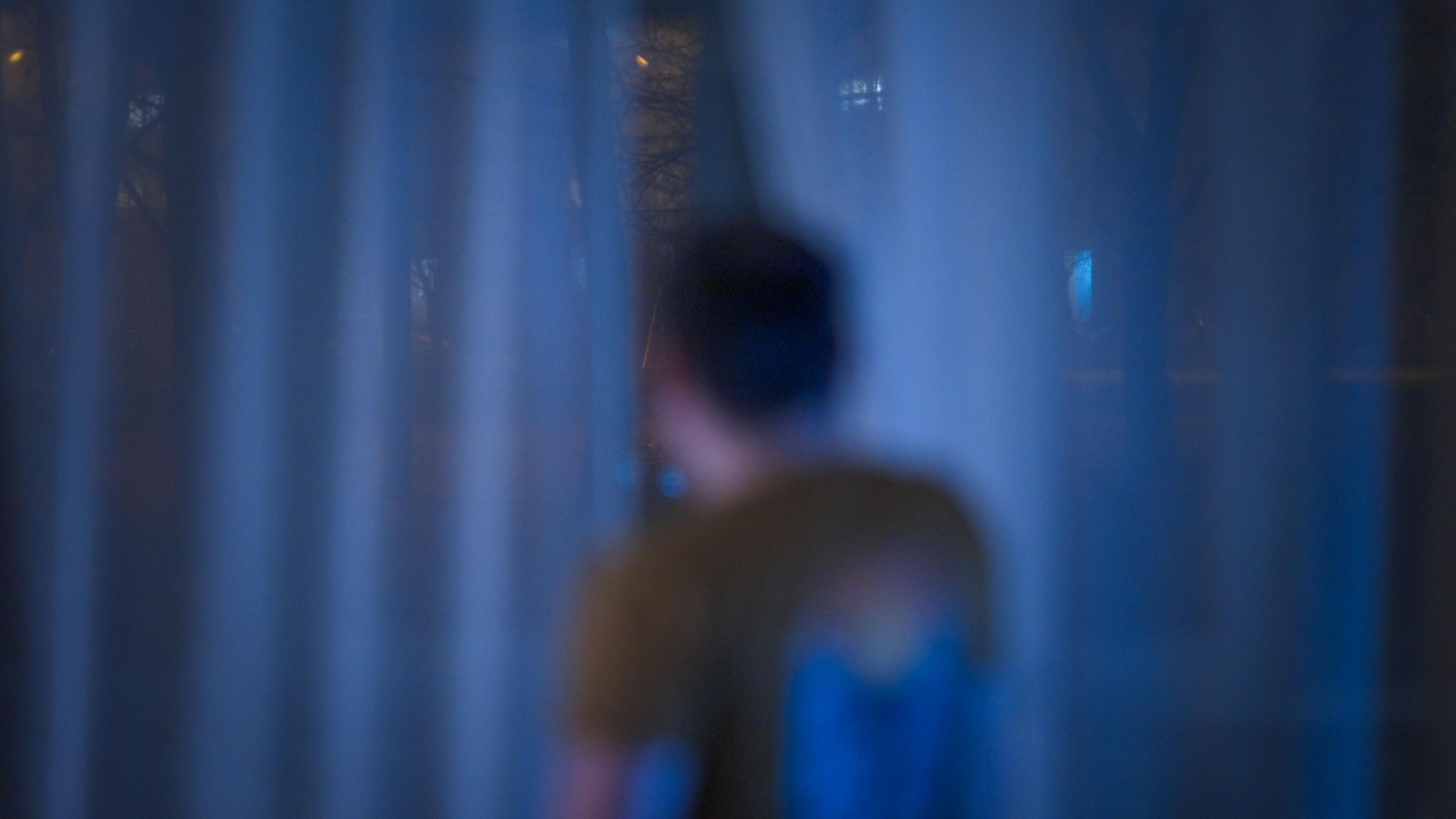 An out of focus image of a man facing away from the camera out of the window through a crack in the curtains.  