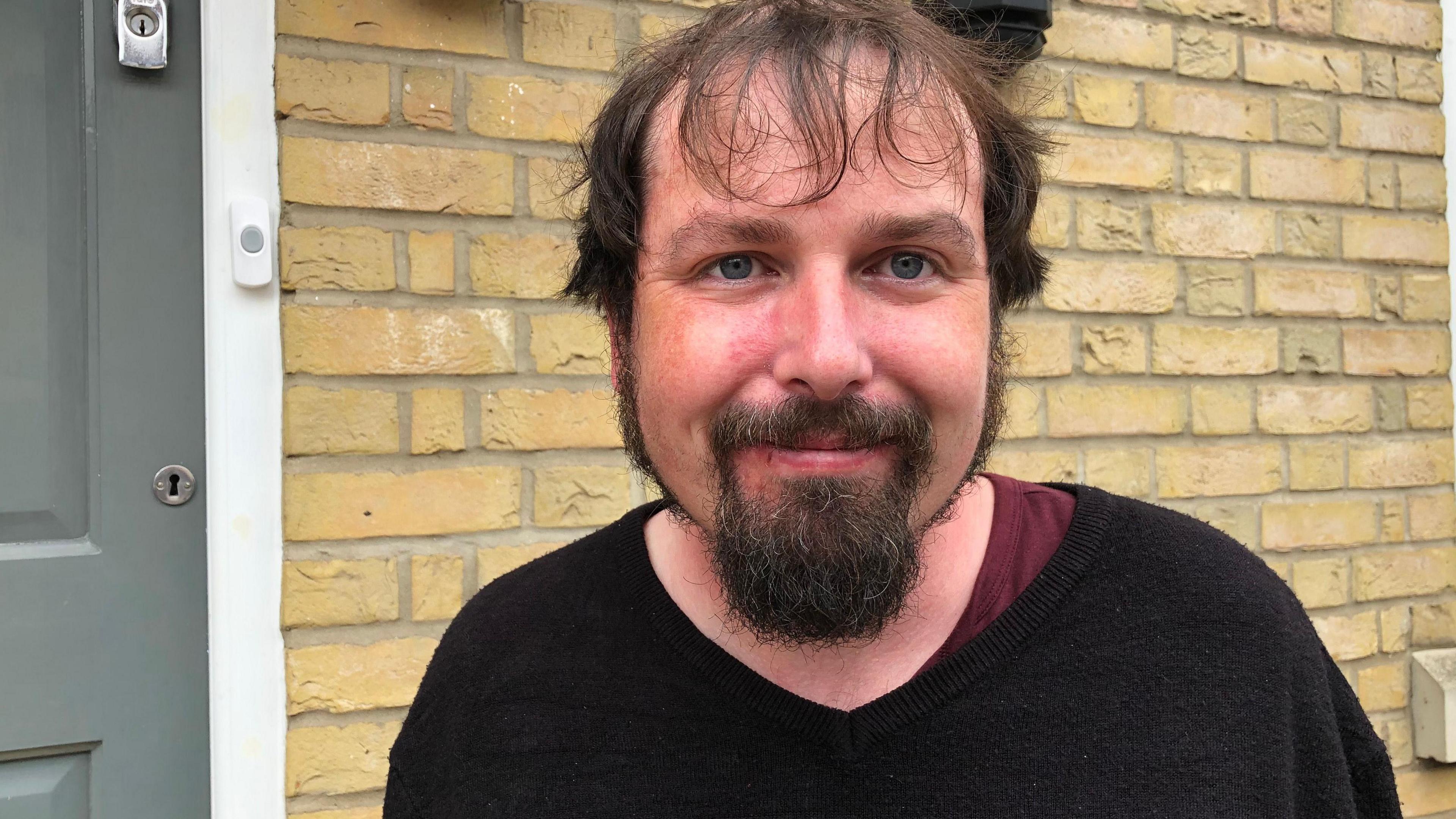 A man with a goatee and wearing a black v-necked jumber over a maroon t-shirt standing against a yellow brick wall