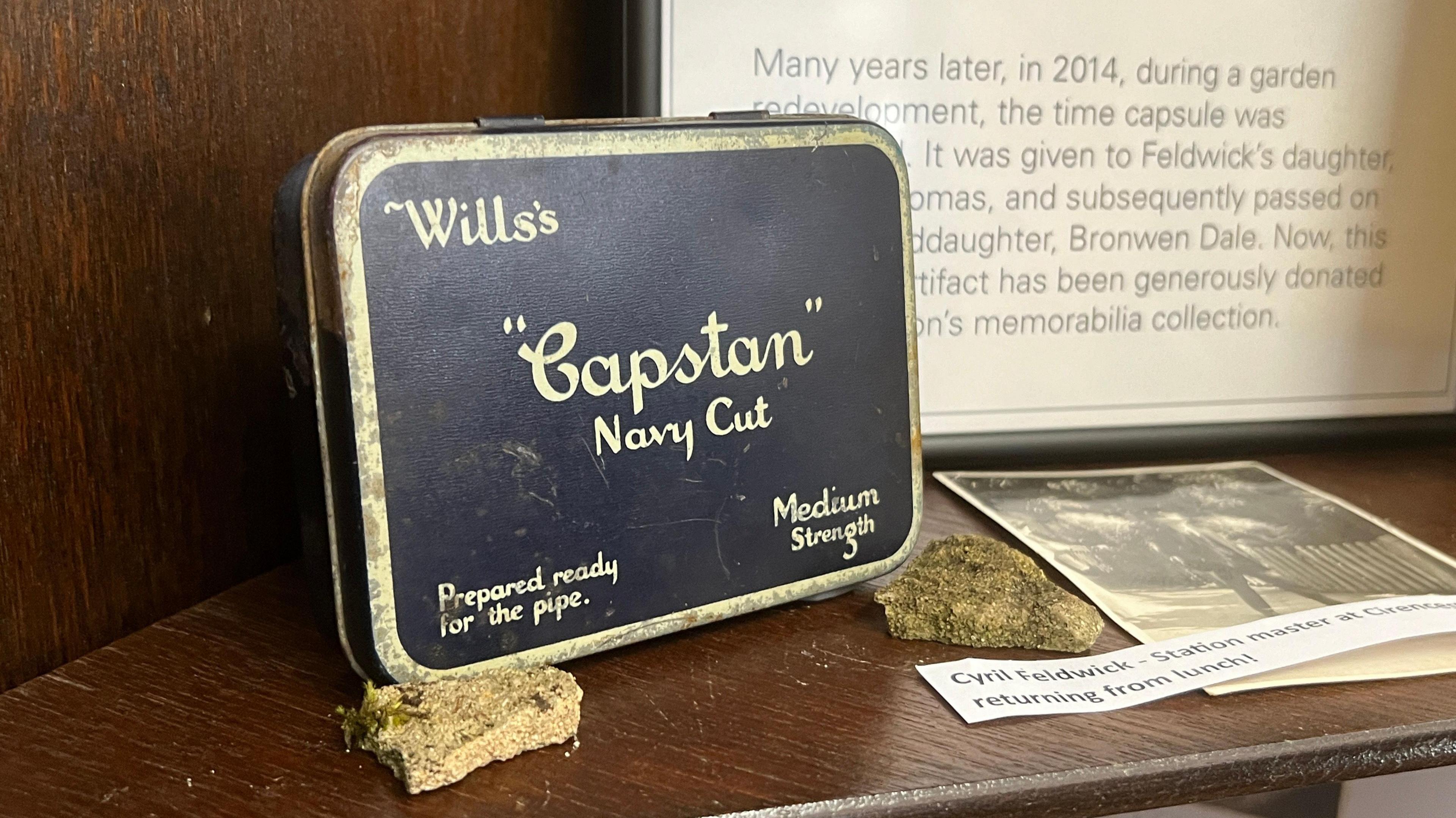 A Wills's "Capston" Navy cut tobacco tin sits on a wooden shelf as part of an exhibition. There are two stone fragments of wall beside it, and a black and white photograph of a man. Behind it, a framed poster, detailing the story behind the time capsule tin, hangs on the wall.