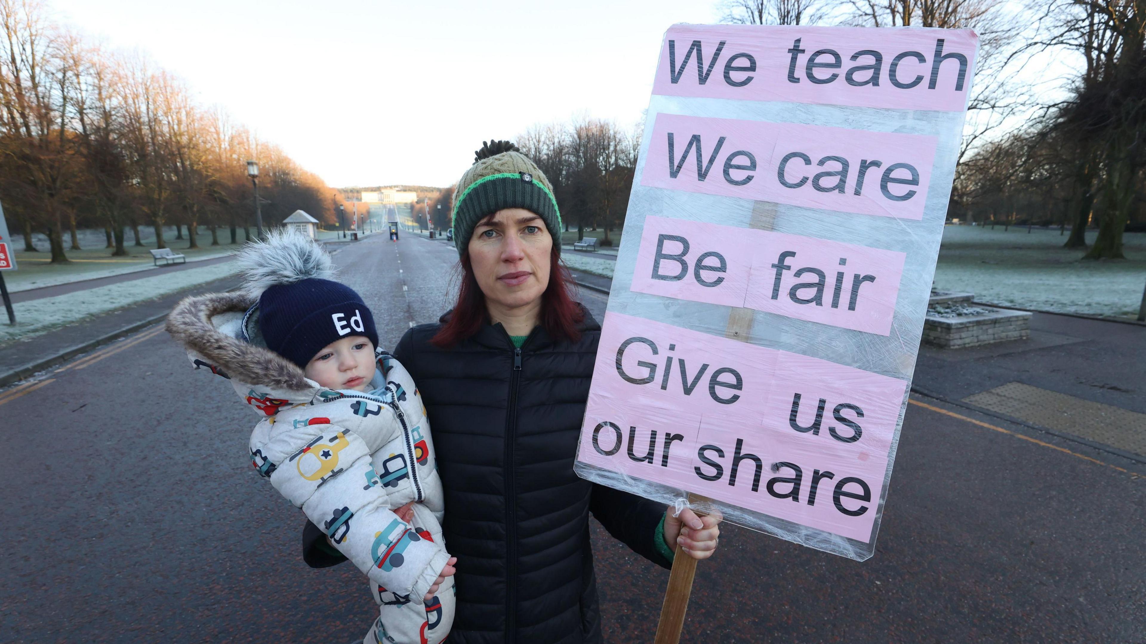 A woman holds a young child in one hand and in the other is holding a large sign which reads 'We teach, we care, be fair, give us our share'. She is wearing a hat and a black jacket, and is standing in the middle of a road which has frosty grass patches on both sides.