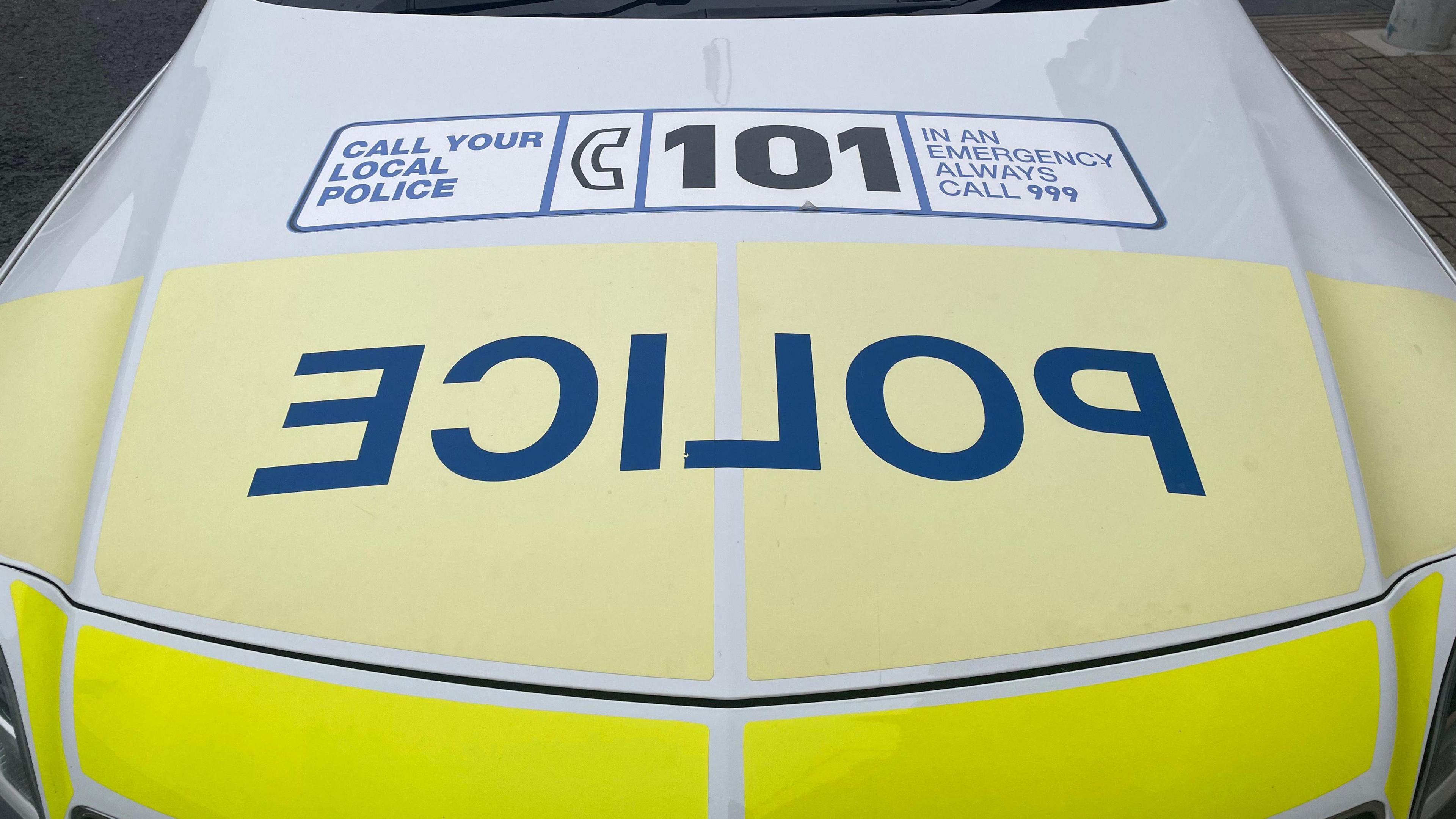 A white and yellow police car with police written in blue block capitals in reverse. The bonnet has a sticker saying to call 101 or 999.