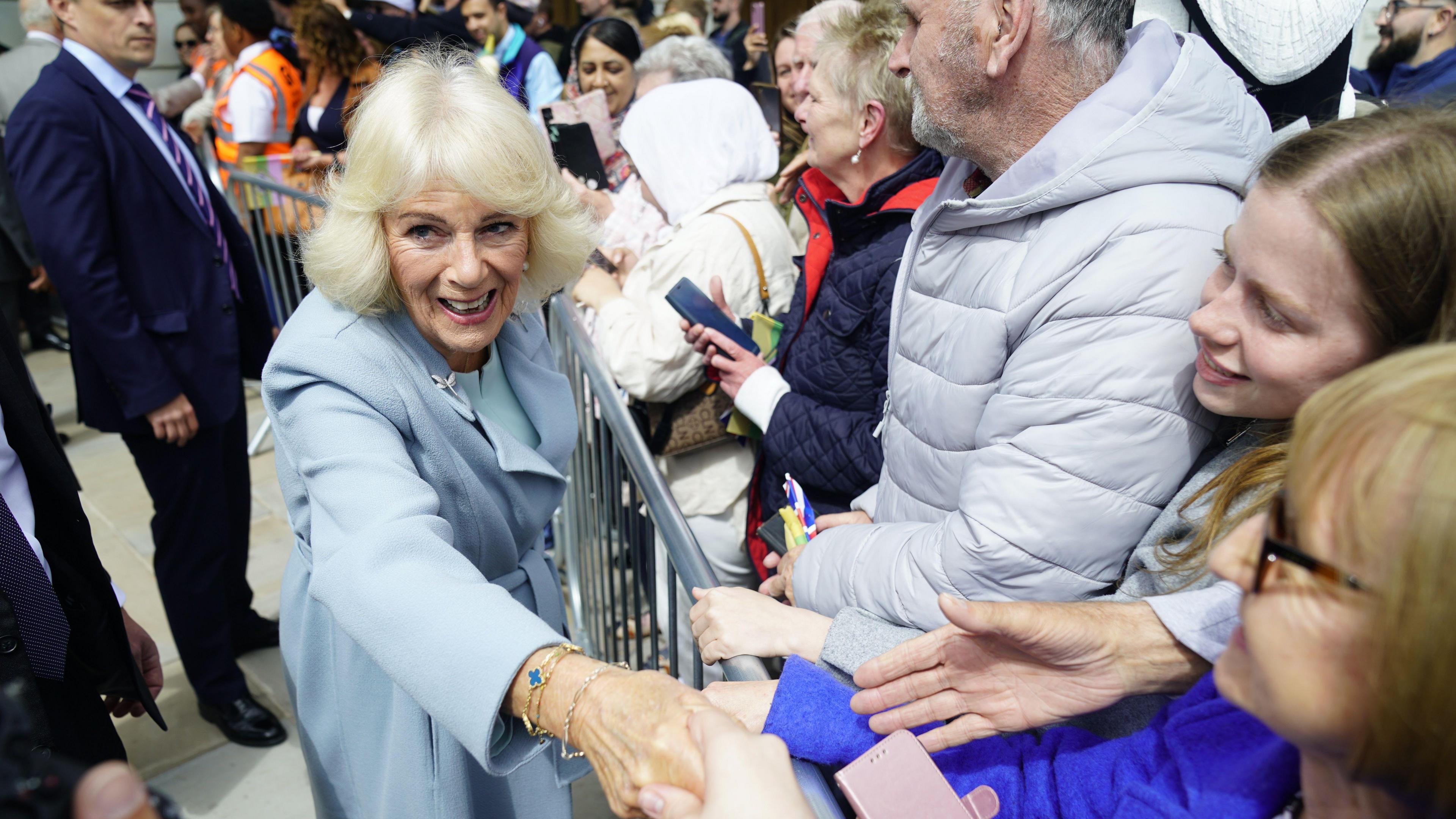 Queen Camilla reaching to shake hands of members of the public over a railing