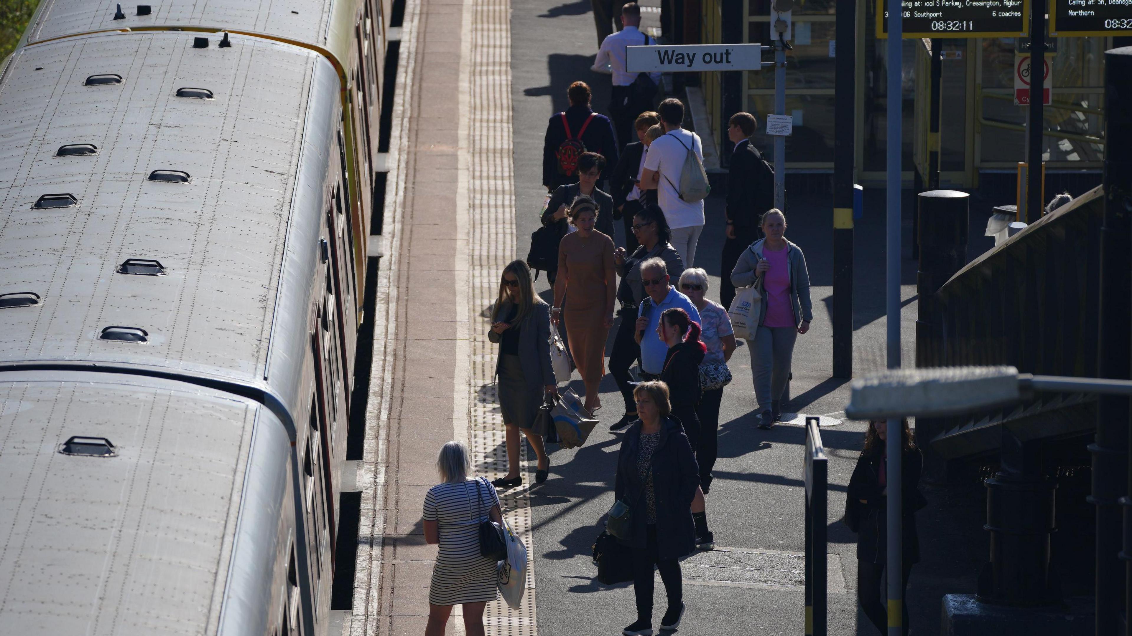 An elevated view of a group of passengers on the platform at Hunts Cross station approaching a train carriage which has just stopped. 