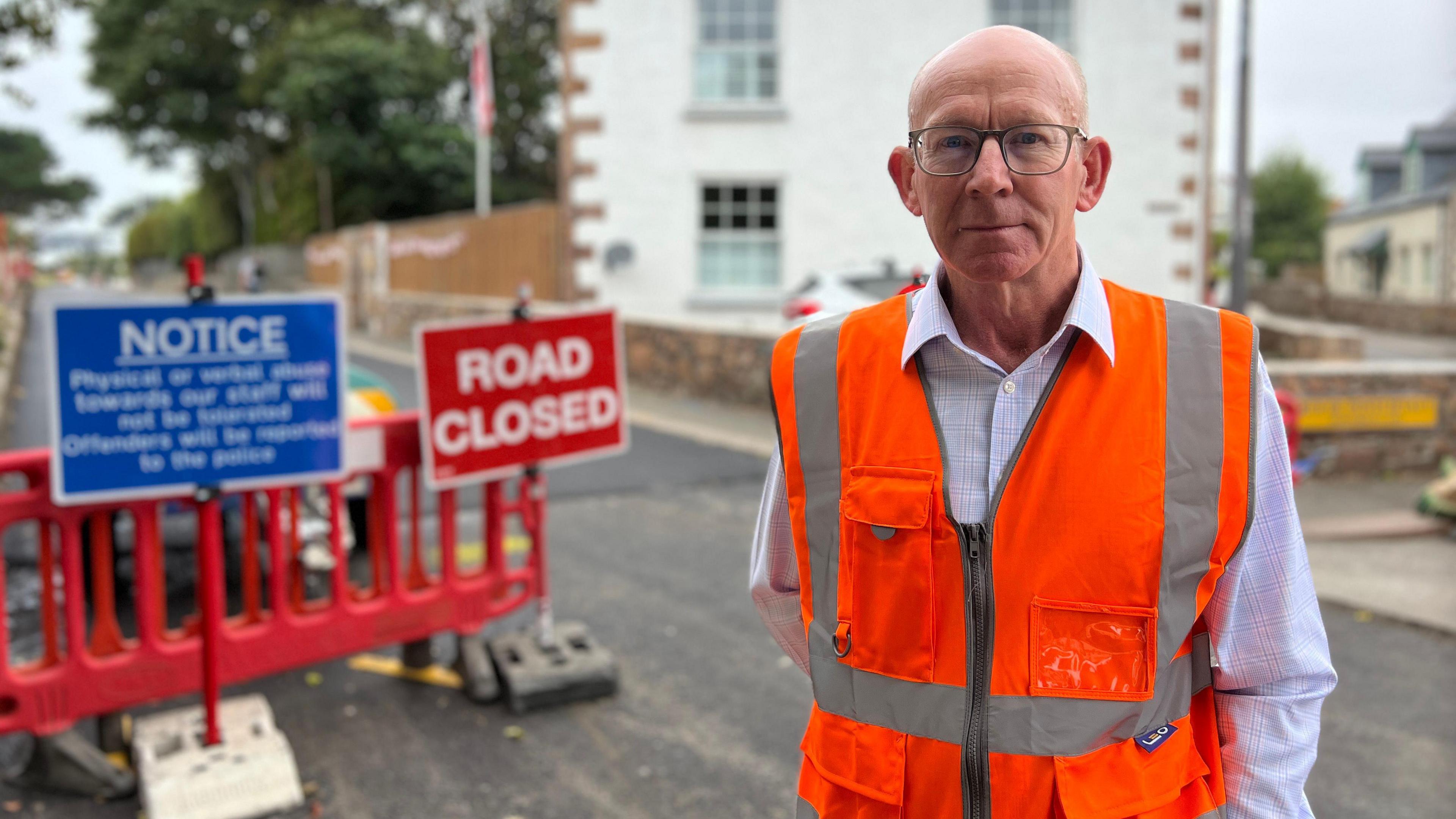 A man wearing a high vis jacket with road closed and work signs in the background on a closed road. Blurred background.