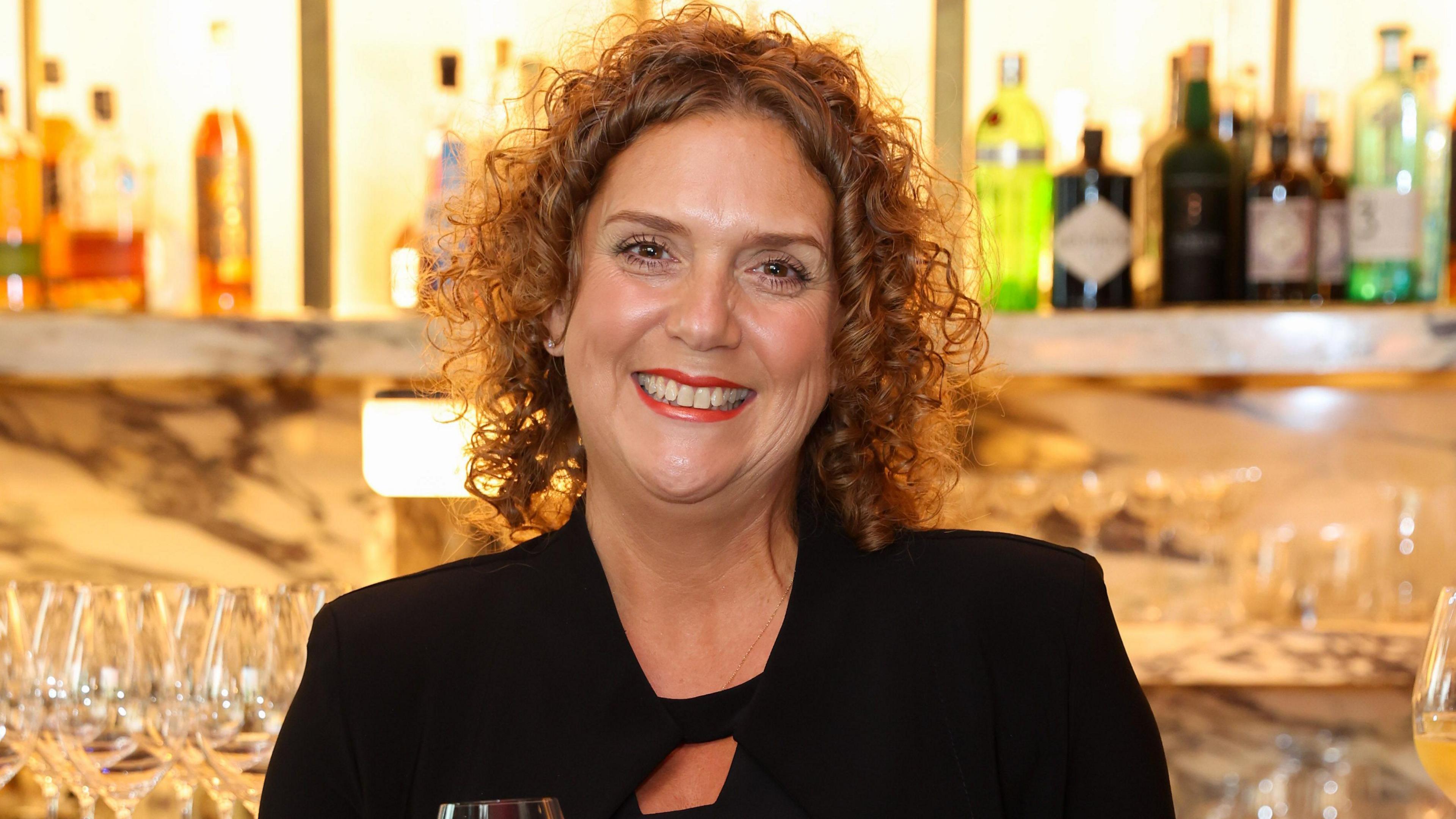 Hannah Ingram-Moore appears in front of a bar, with glasses and bottles behind her. She wears a black jacket. Her hair is curly and she is wearing red lipstick.