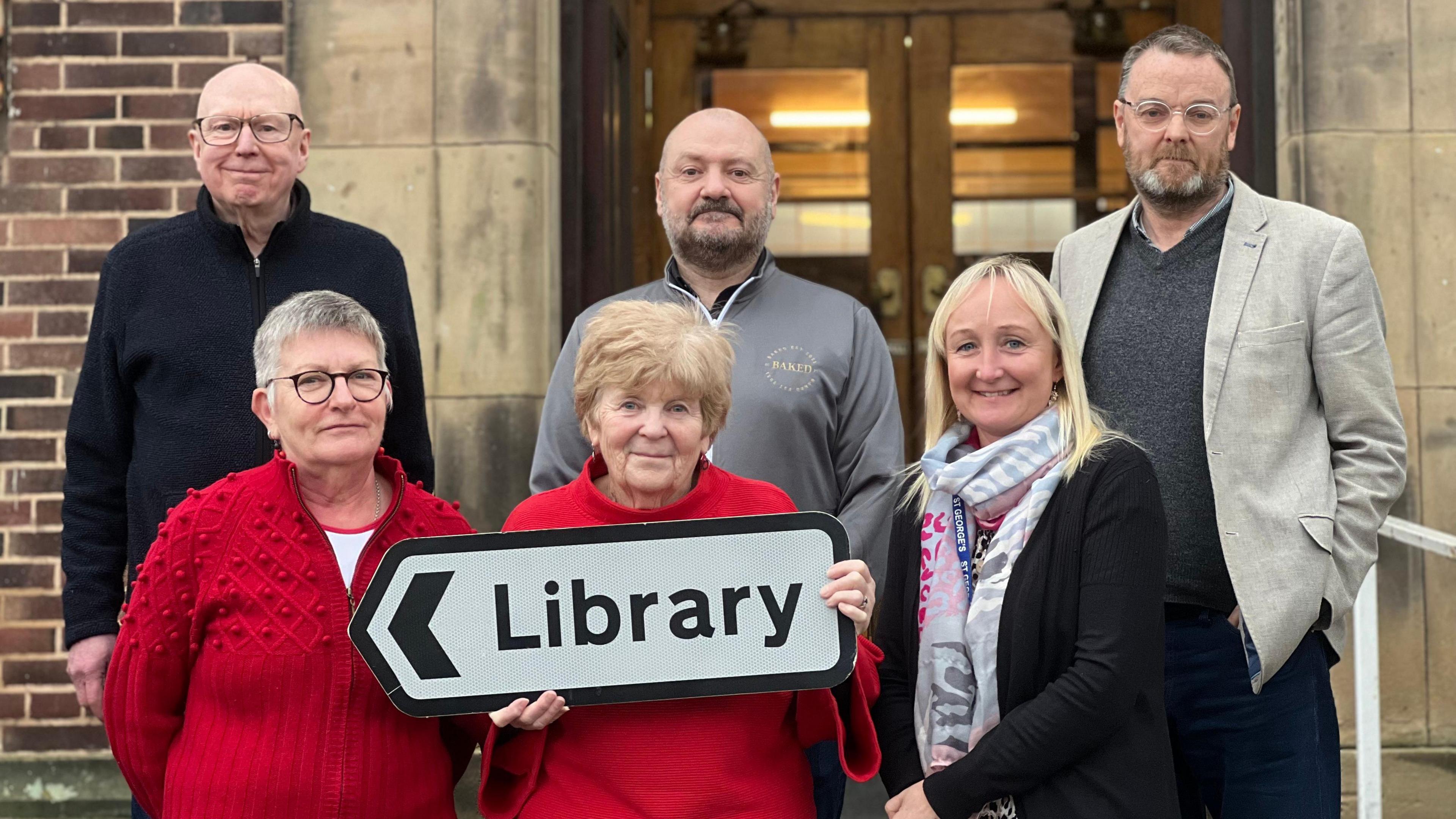 Three men and two women stand outside library doors. A woman wearing red holds a street sign which reads 'library' on it 