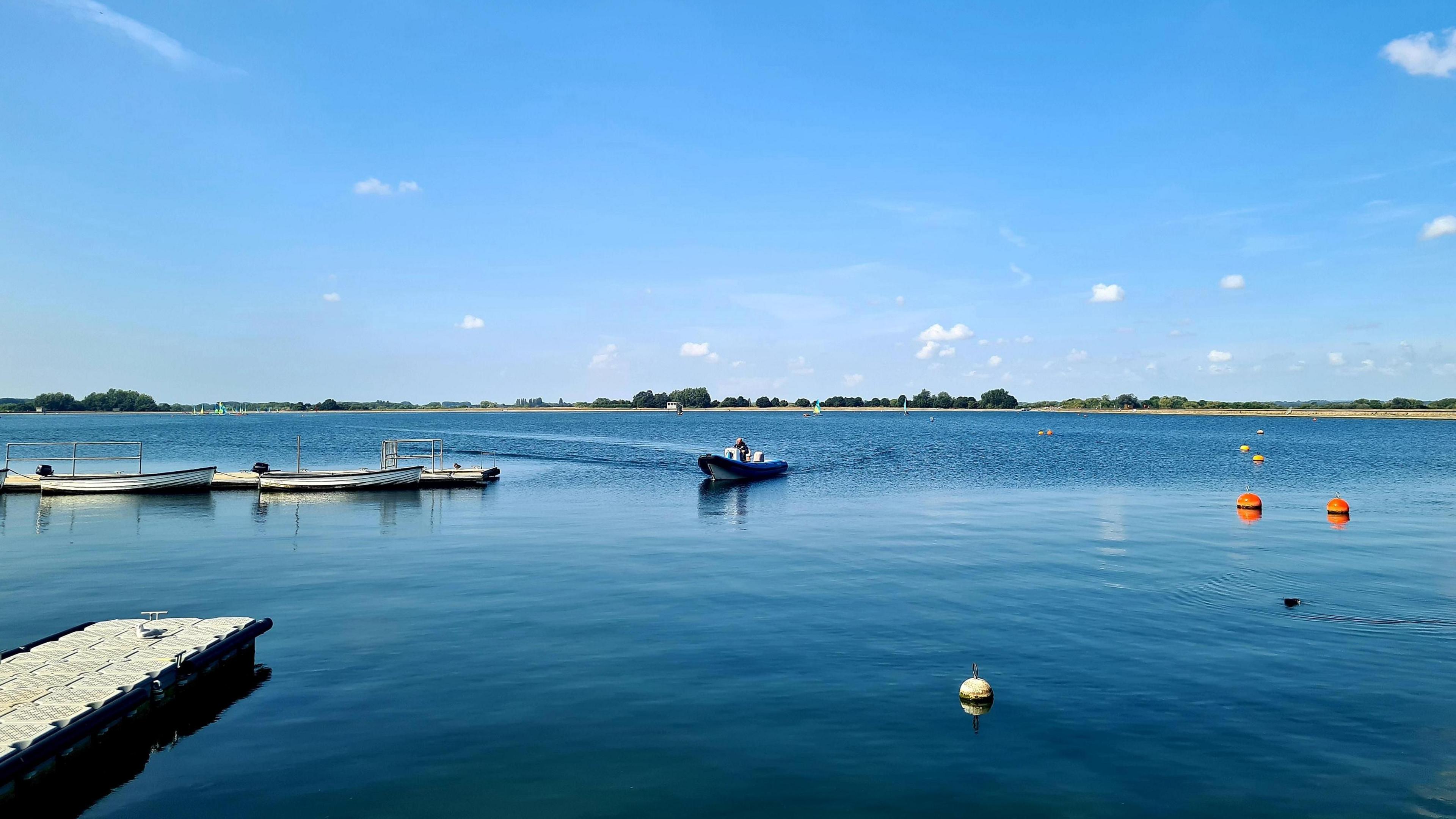 A RIB can be seen on the water which also includes buoys, other small boats and a pontoon. It is a beautiful sunny day with a bright blue sky and a few wispy white clouds.