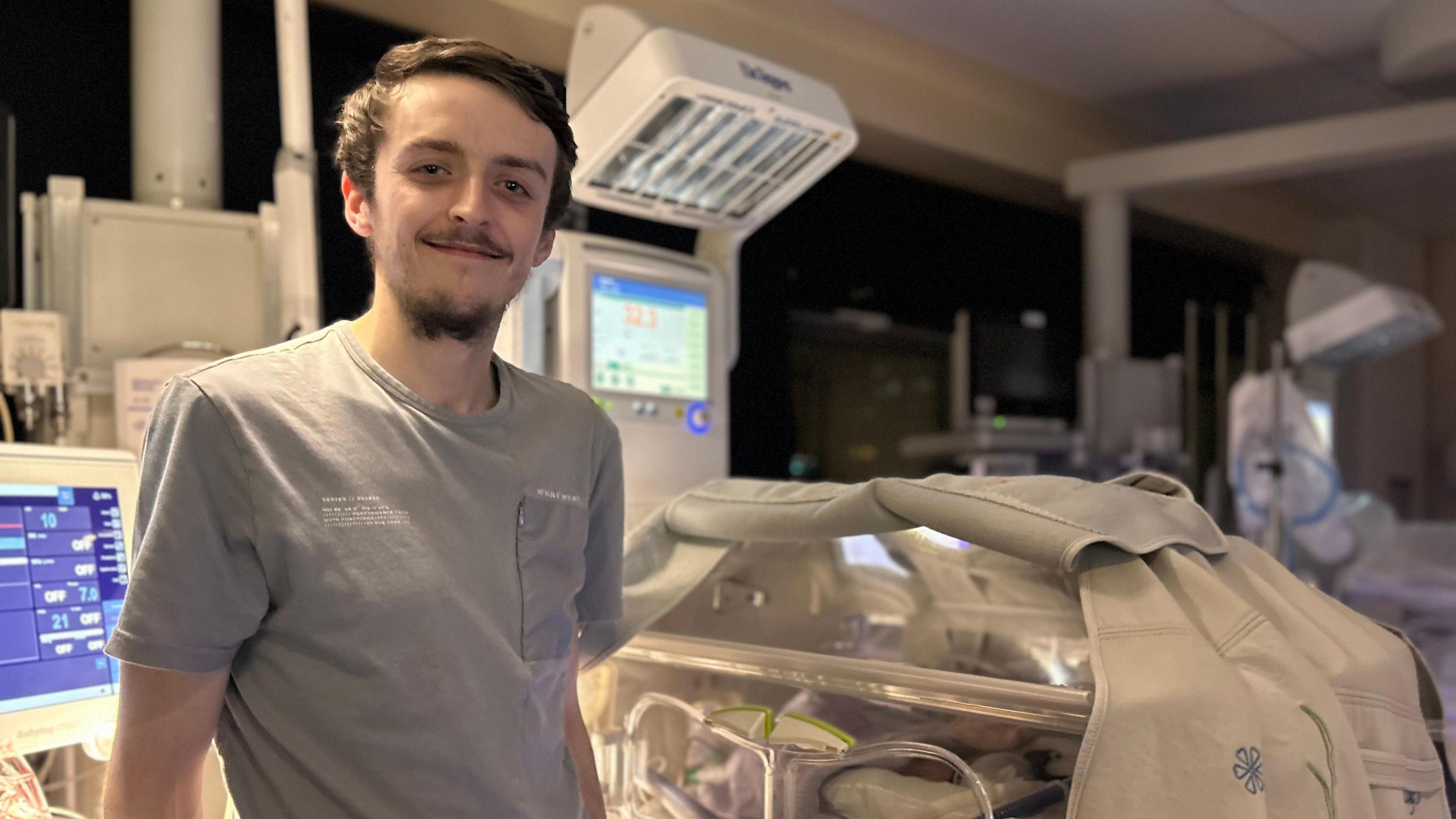 Rob wears a grey t-shirt. He is stood in front of a wall of medical monitoring equipment and is next to an intensive care crib, with one of his daughters inside.