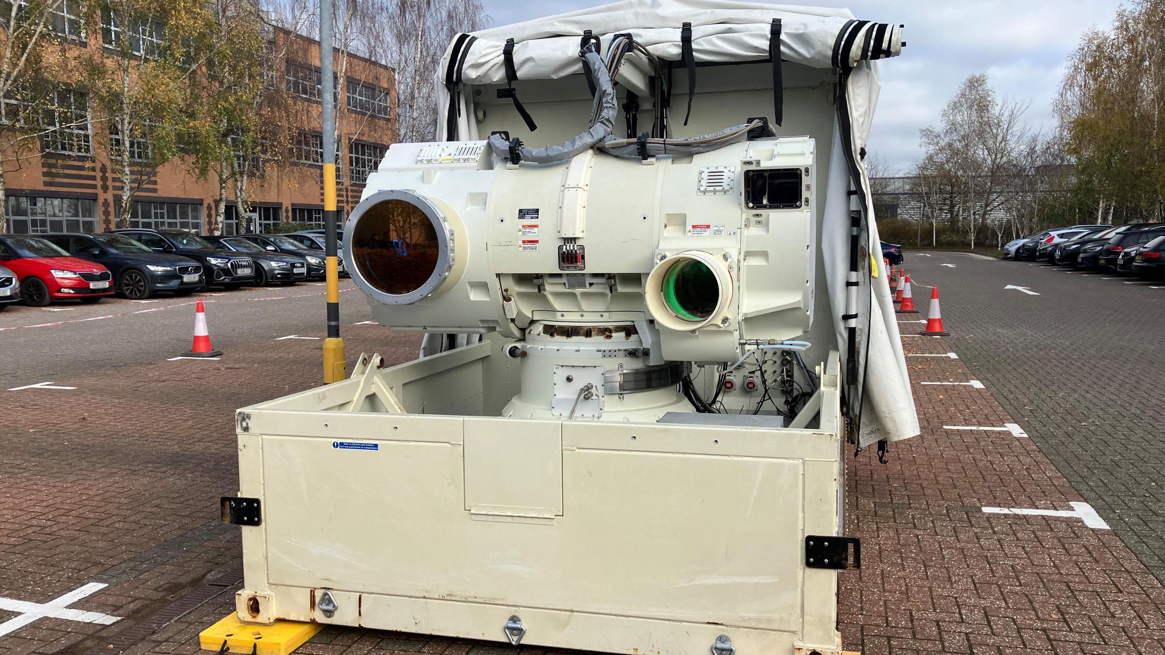 A large white military-grade laser system mounted on a platform, featuring two prominent circular lenses and multiple cables.