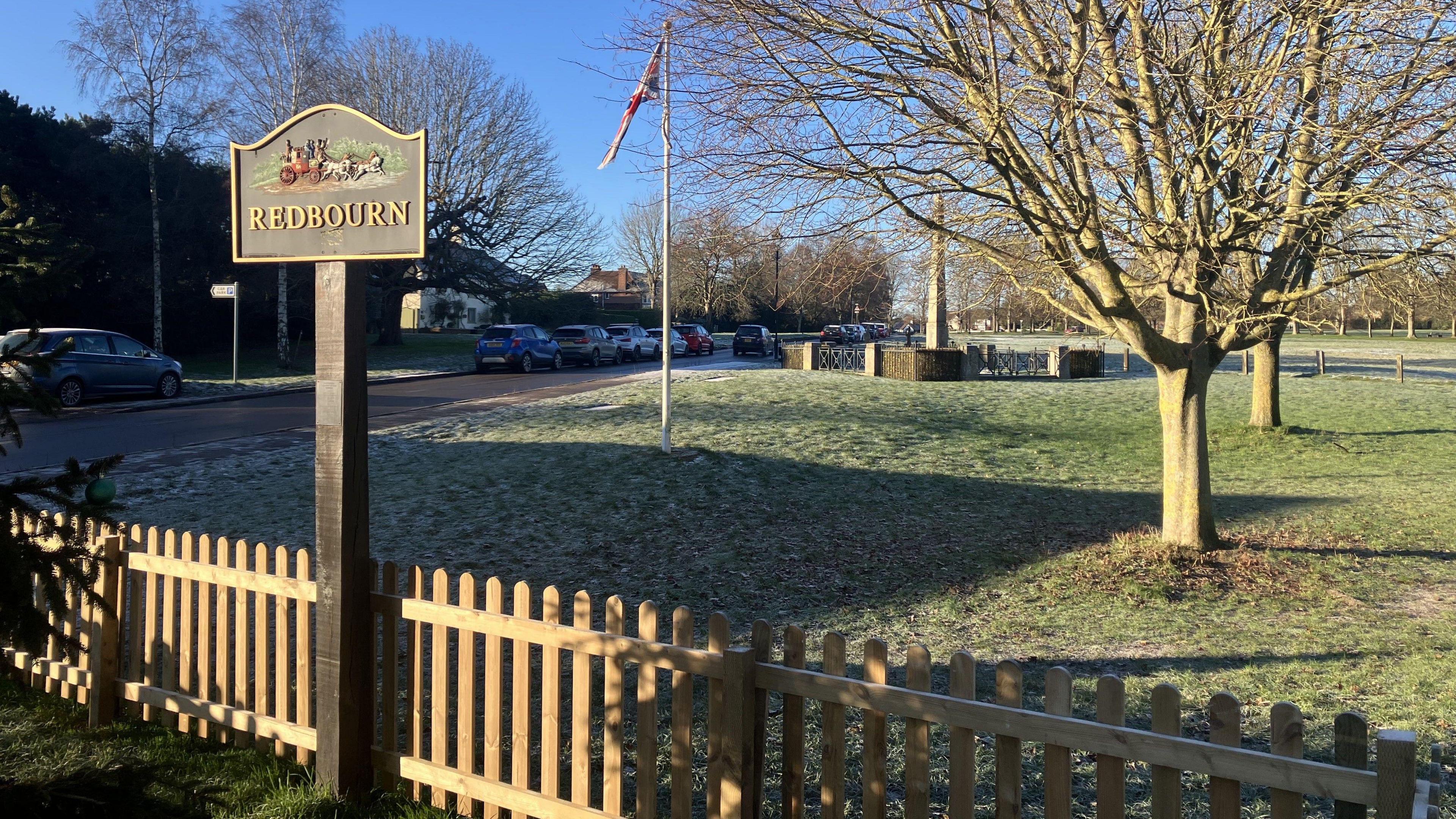 A picture taken in Redbourn village in Hertfordshire. There is a sign that says Redbourn at the front of the picture, in the middle a union flag at half mast plus a war memorial. On the right of the image there are two trees and the sky is blue in the background.