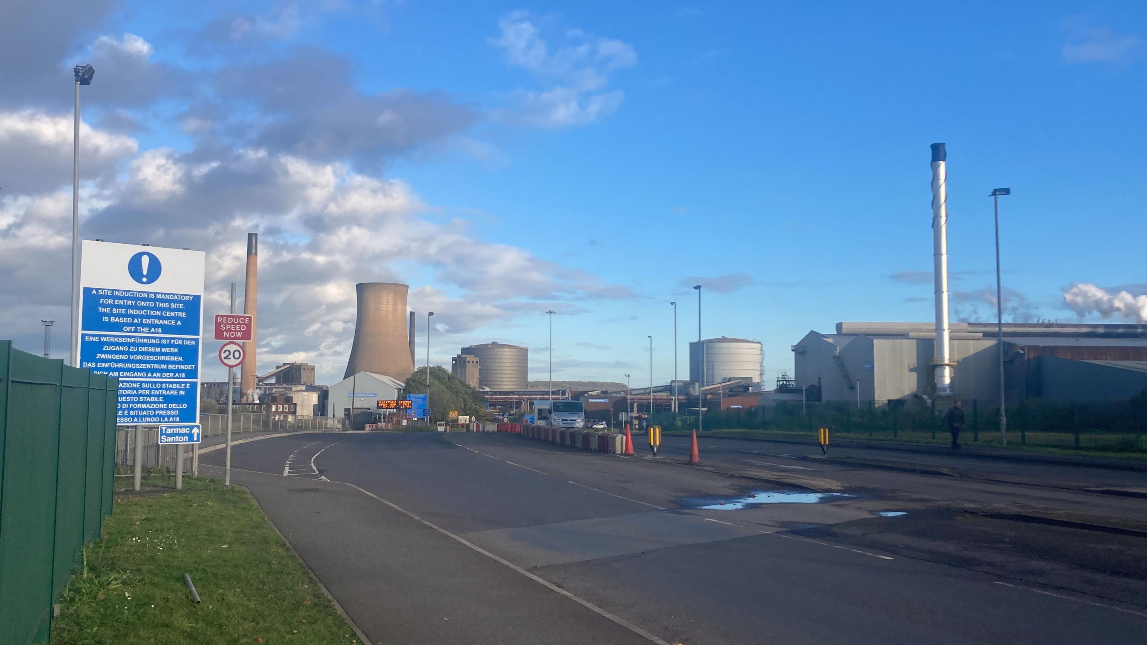 A view of Scunthorpe steelworks. A wide road runs towards cooling towers, metal chimneys and large buildings. To the left is a green fence and a large sign with white text on a blue background.