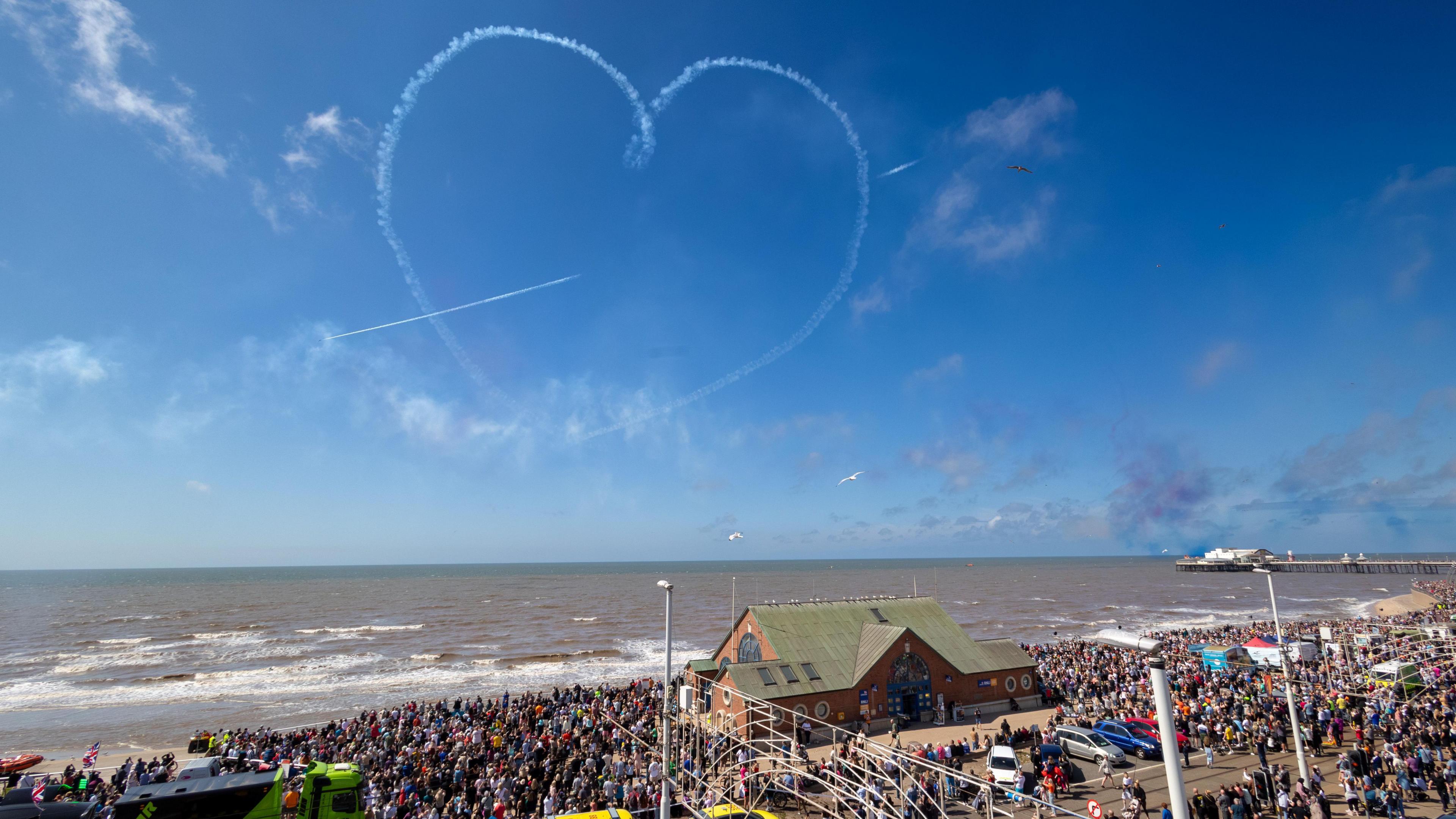 A photo of a sunny day at Blackpool beach. thousands of people can be seen in the crowd on the seafront. In the blue skies above, a plane has drawn a heart in the sky. There's a building in the middle of the beach, and a pier in the distance