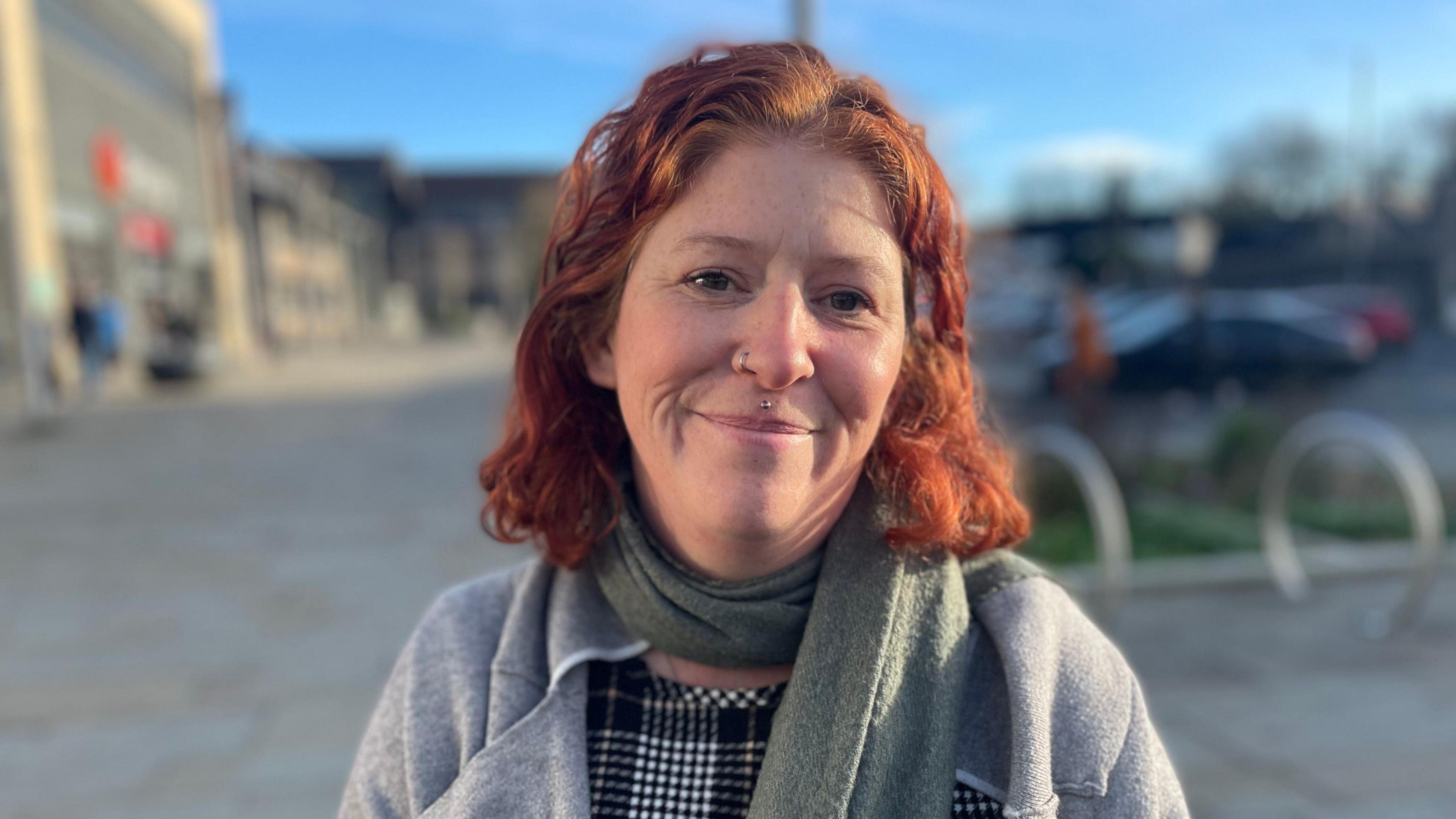 Menyw gyda gwallt coch i'w hysgwyddau, yn gwisgo sgarff werdd a chot lwyd yn sefyll ar stryd yng nghanol y dref