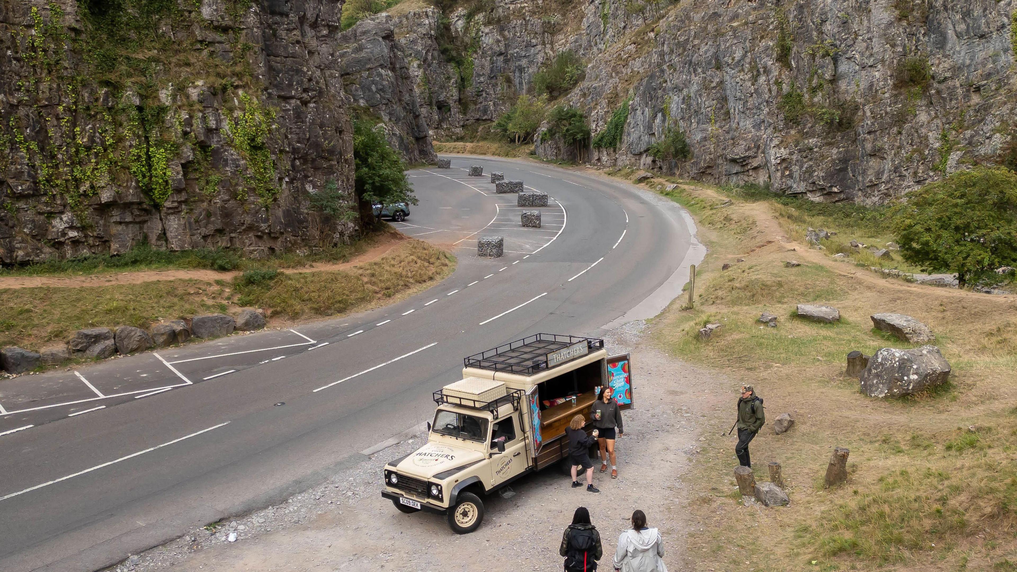 An aerial shot of a road going through Cheddar gorge, with cliffs on either side. A truck is parked by the side of the road selling cider, people are approaching it.