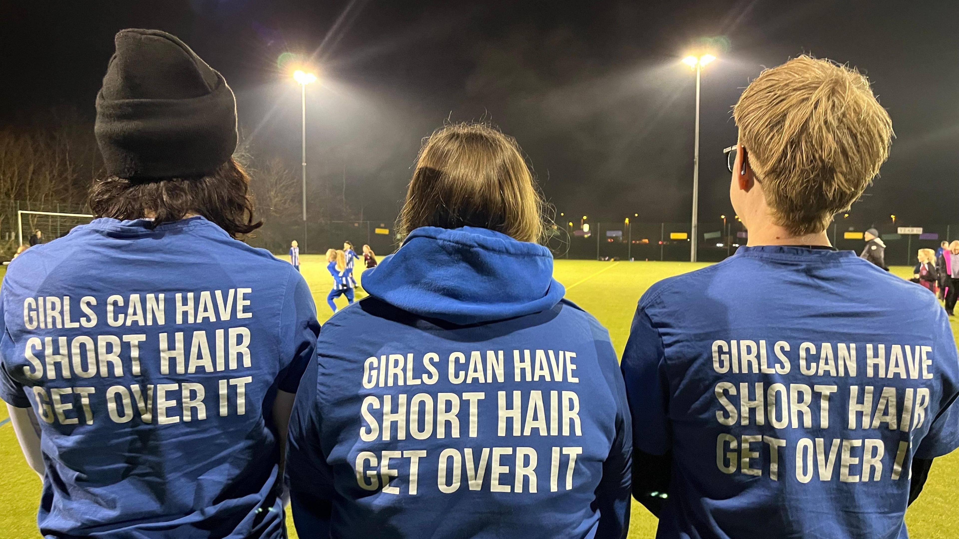 Three women have their backs to the camera to show their blue t-shirts with the words "Girls can have short hair - Get over it".
They are standing in front of a flood-lit football pitch with young girls playing football.

 