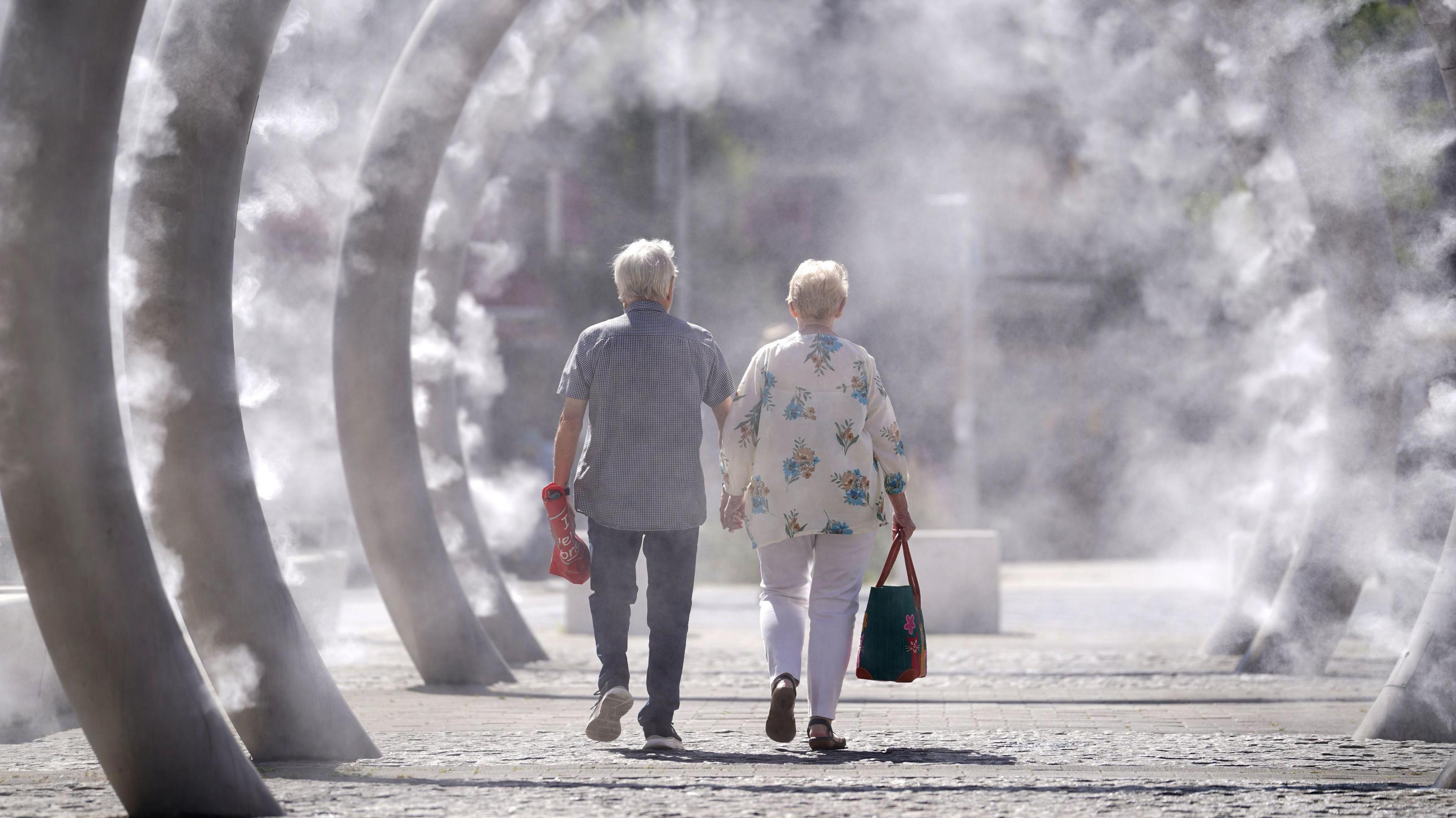 A man and a woman - both with silver hair - are walking inside a row of ring metal structures that are spraying mist on a very sunny day. A man is wearing a checkered blue shirt with short sleeves, jeans and trainers and a woman in white trousers and a loose-fitting shirt.