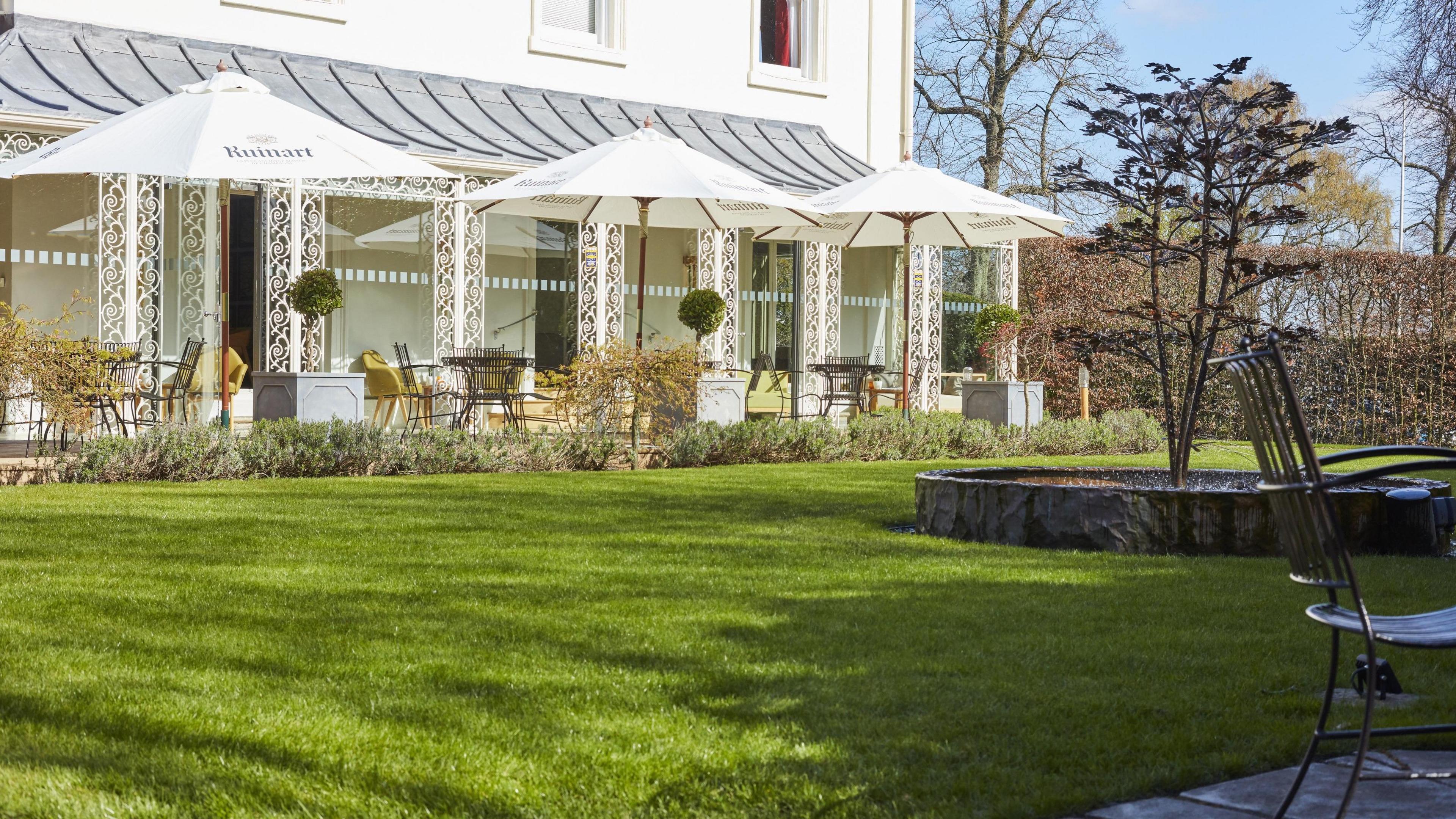 The garden at Simpsons restaurant in Birmingham with a lawn, a patio, tables and chairs sheltered by square parasols and plants in containers.