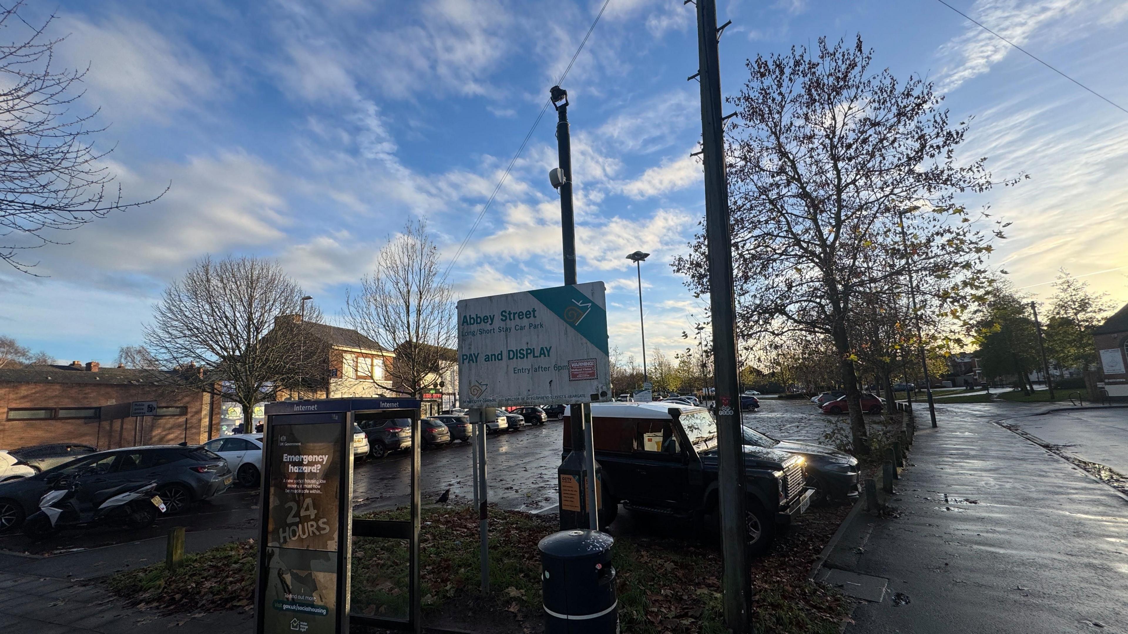 In low sun a surface car park set between roads with parked cars and spaces stretching into the middle distance. A dirty sign reading "Abbey Street Pay and Display" and a phone box in the foreground.