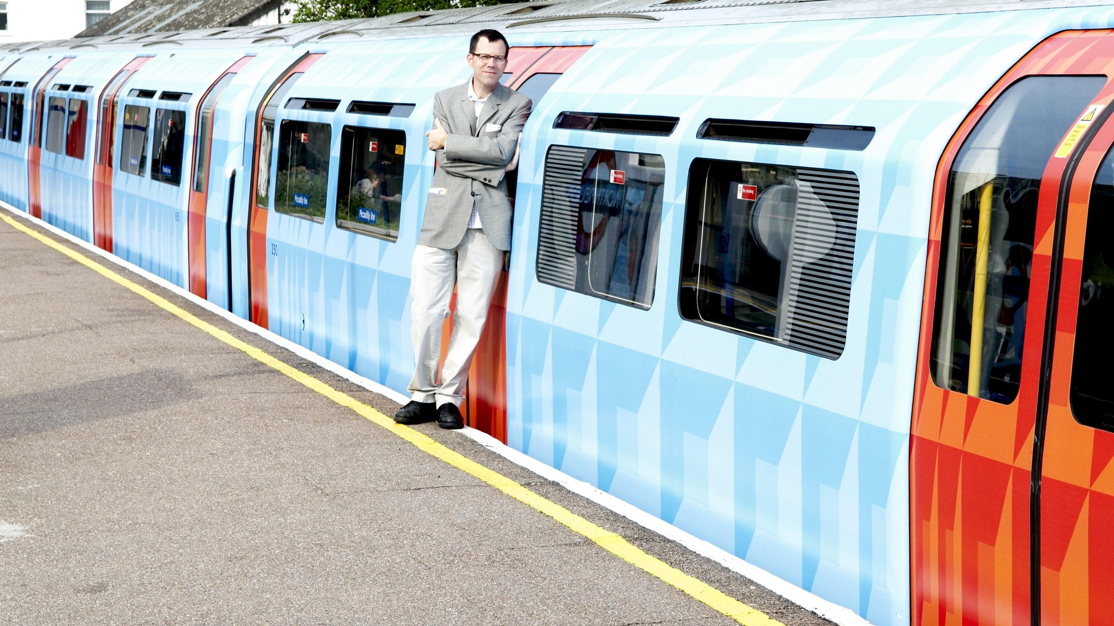 A man in a light grey jacket and white trousers leans against a tube train at a platform. The exterior of the train is wrapped in a blue and red geometric design. 