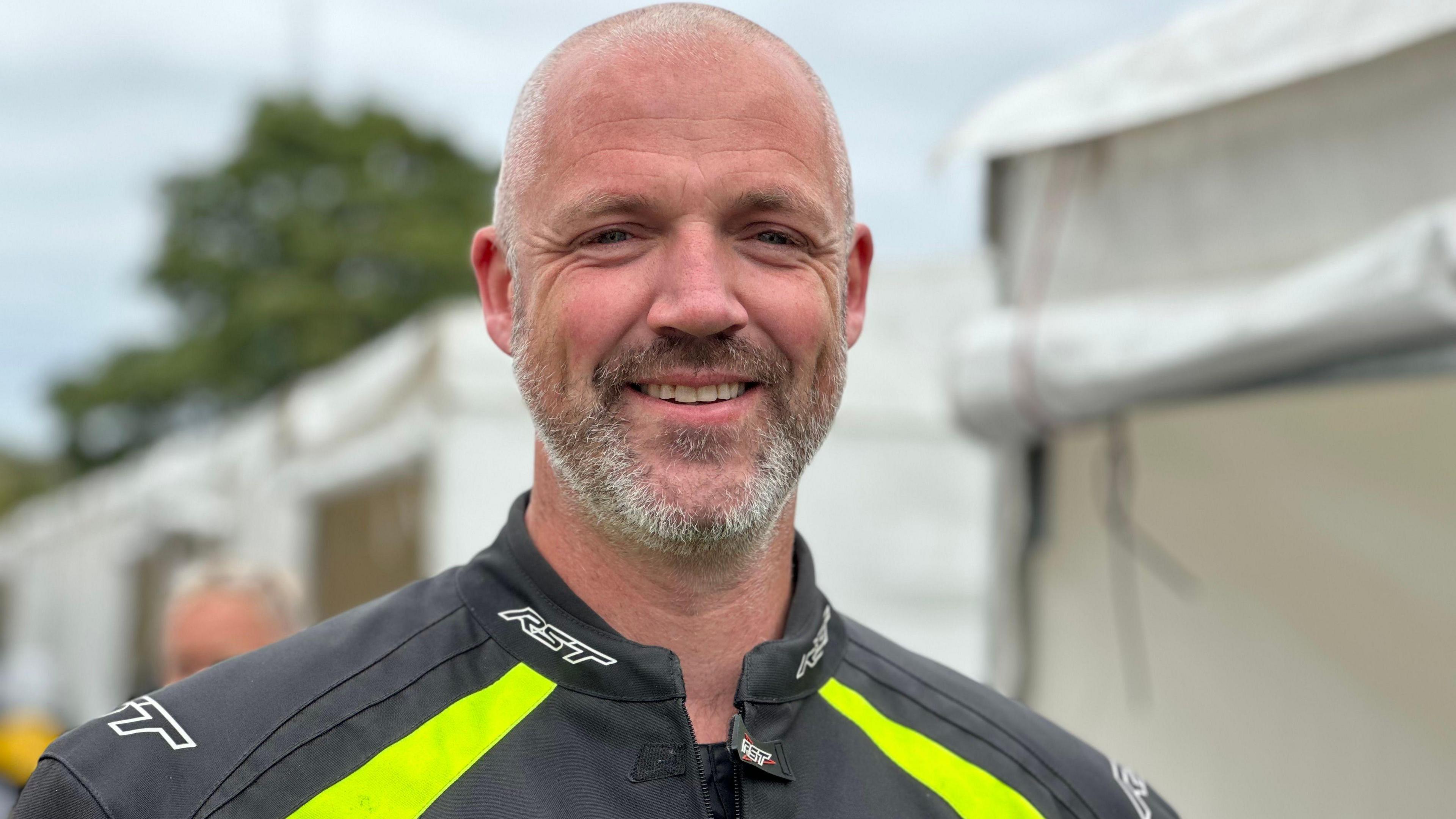John Eastham has short grey hair and a beard, he wears a black jacket with a fluorescent yellow stripe.
