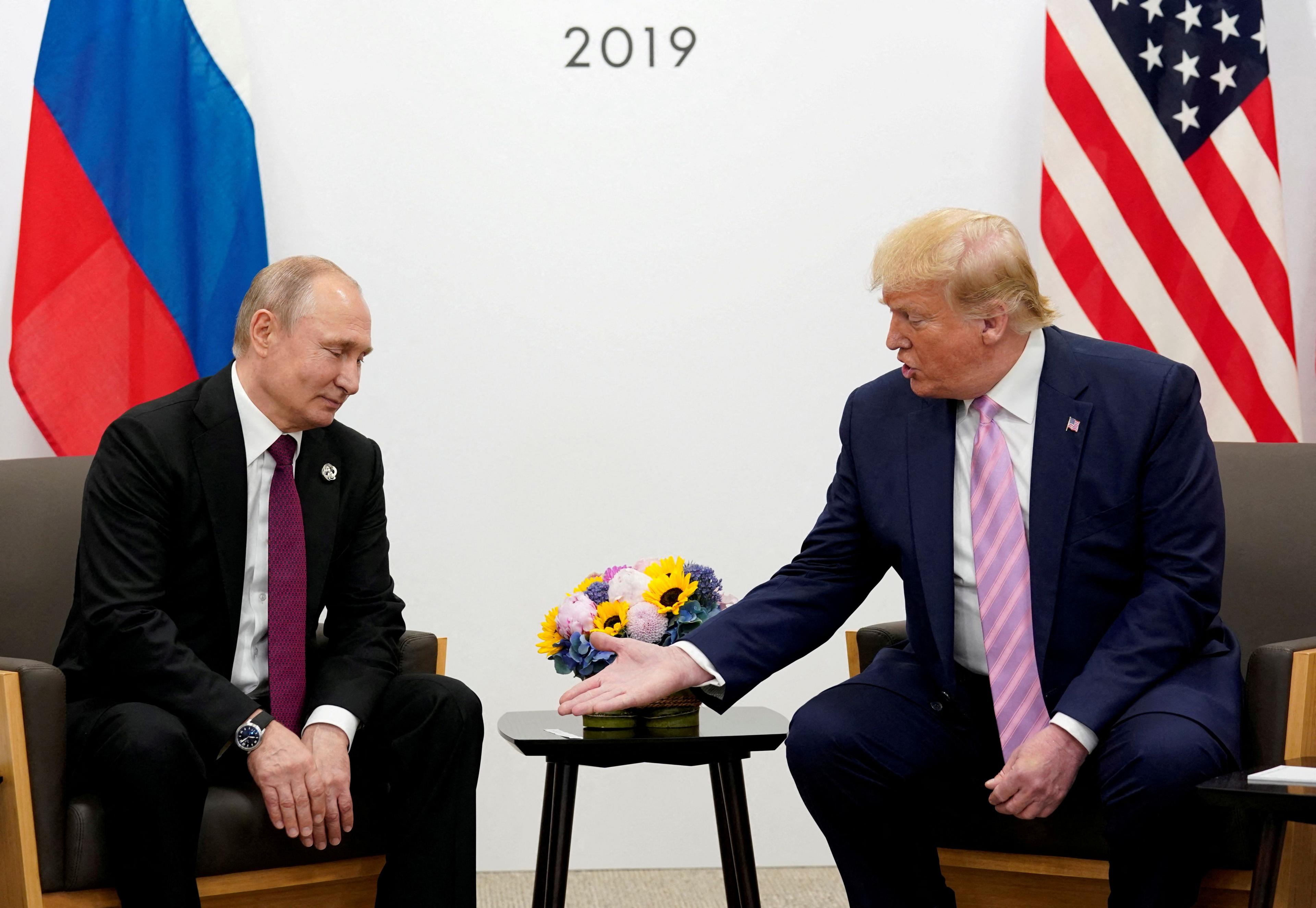 Donald Trump gestures towards Russian President Vladimir Putin at the G20 leaders summit in Japan. Both men are wearing suits and are sitting in dark coloured chairs in front of their country's respective flags. 