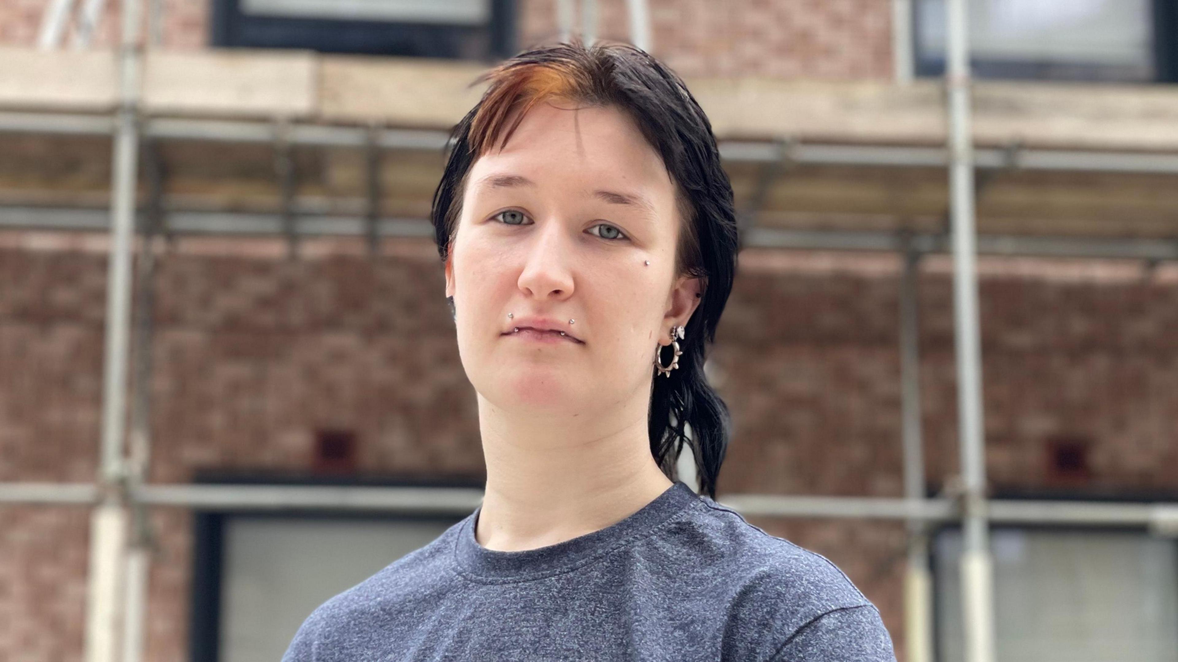 Person with dark hair and piercings in ears and face stands outside some flats wrapped in scaffolding