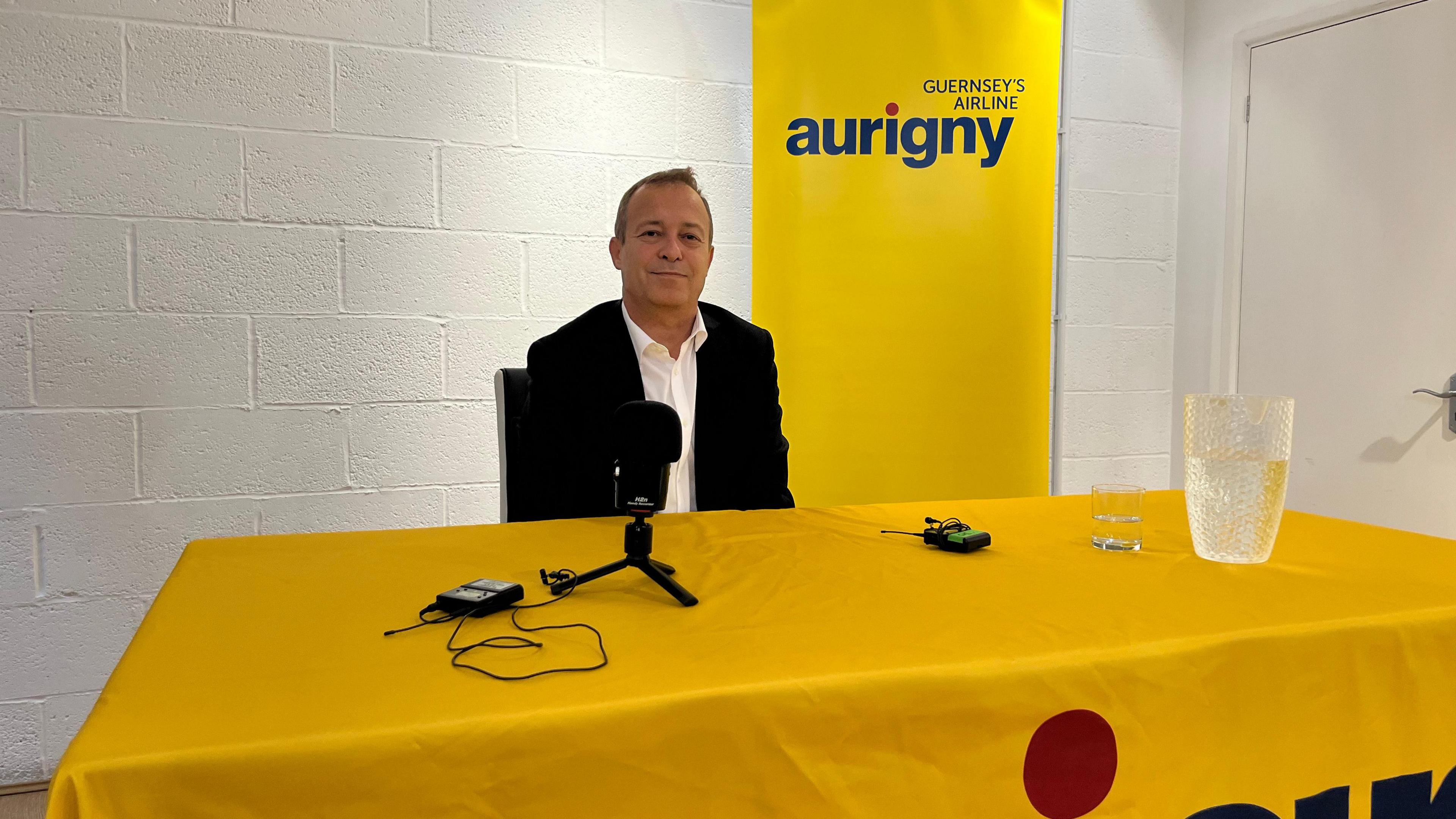 A man sat behind a table with a yellow tablecoth and a sign saying Aurigny behind him. The man is wearing a suit with a white shirt and in front of him on the table is a microphone on a small stand. 