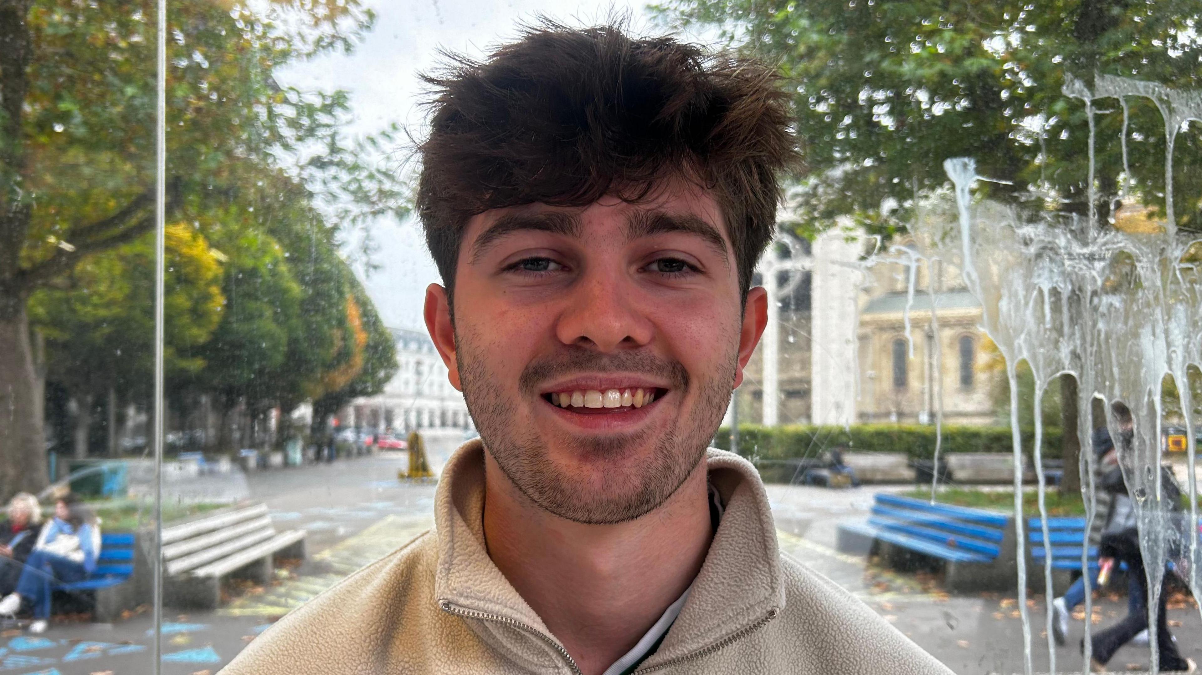 A person with short brown hair smiles at the camera. They are wearing a beige jacket, and appearing to be in a city. In the background are a number of trees.