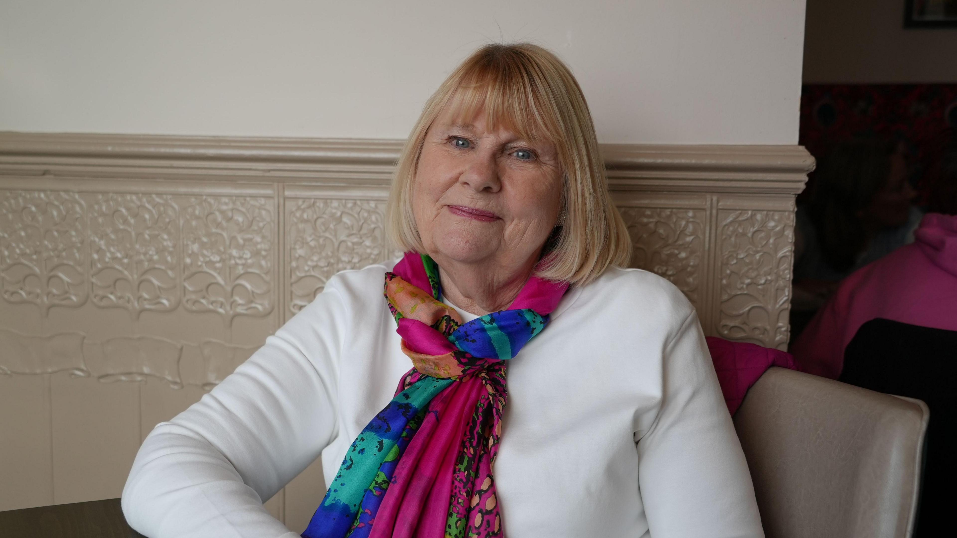 Gloria Reynolds looking into the camera. She is wearing a white top and a colourful scarf. She is sitting in a chair. 