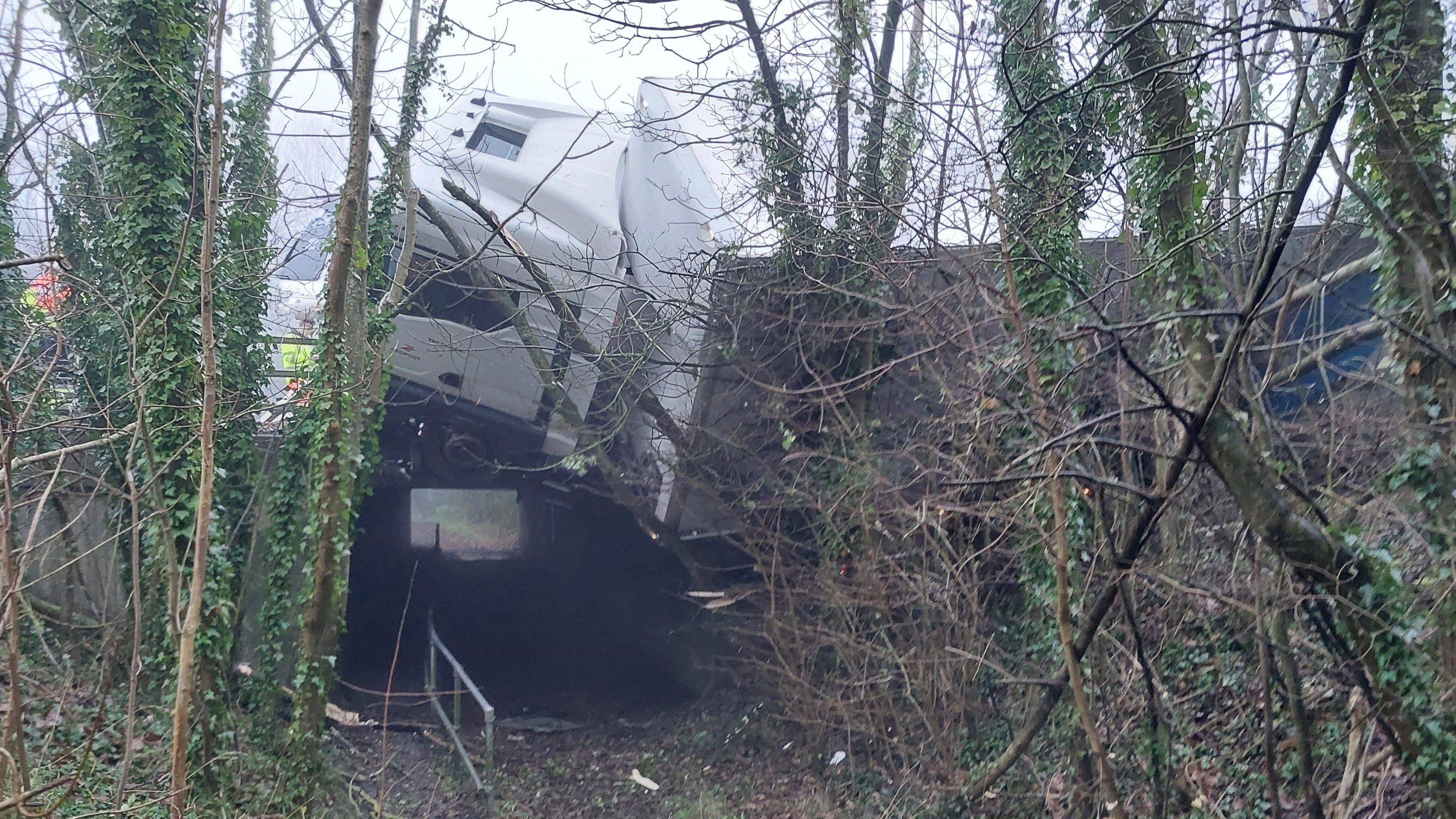 A lorry hanging off a road. Below is a footpath surrounded by trees.