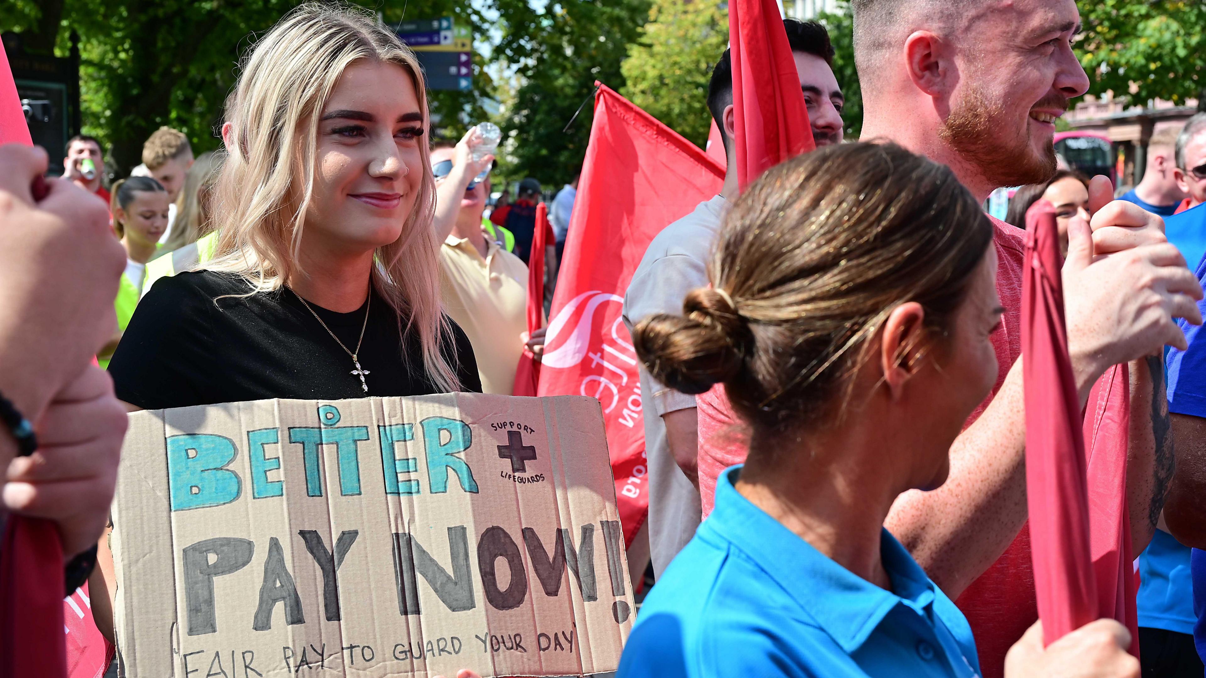 A young woman with long blonde hair holds a brown cardboard sign which says 'better pay now, fair pay to guard your day'. She is wearing a black t-shirt and a silver cross necklace. There are other men and women surrounding her holding red union flags. They are wearing red, blue, white and other coloured clothing.
