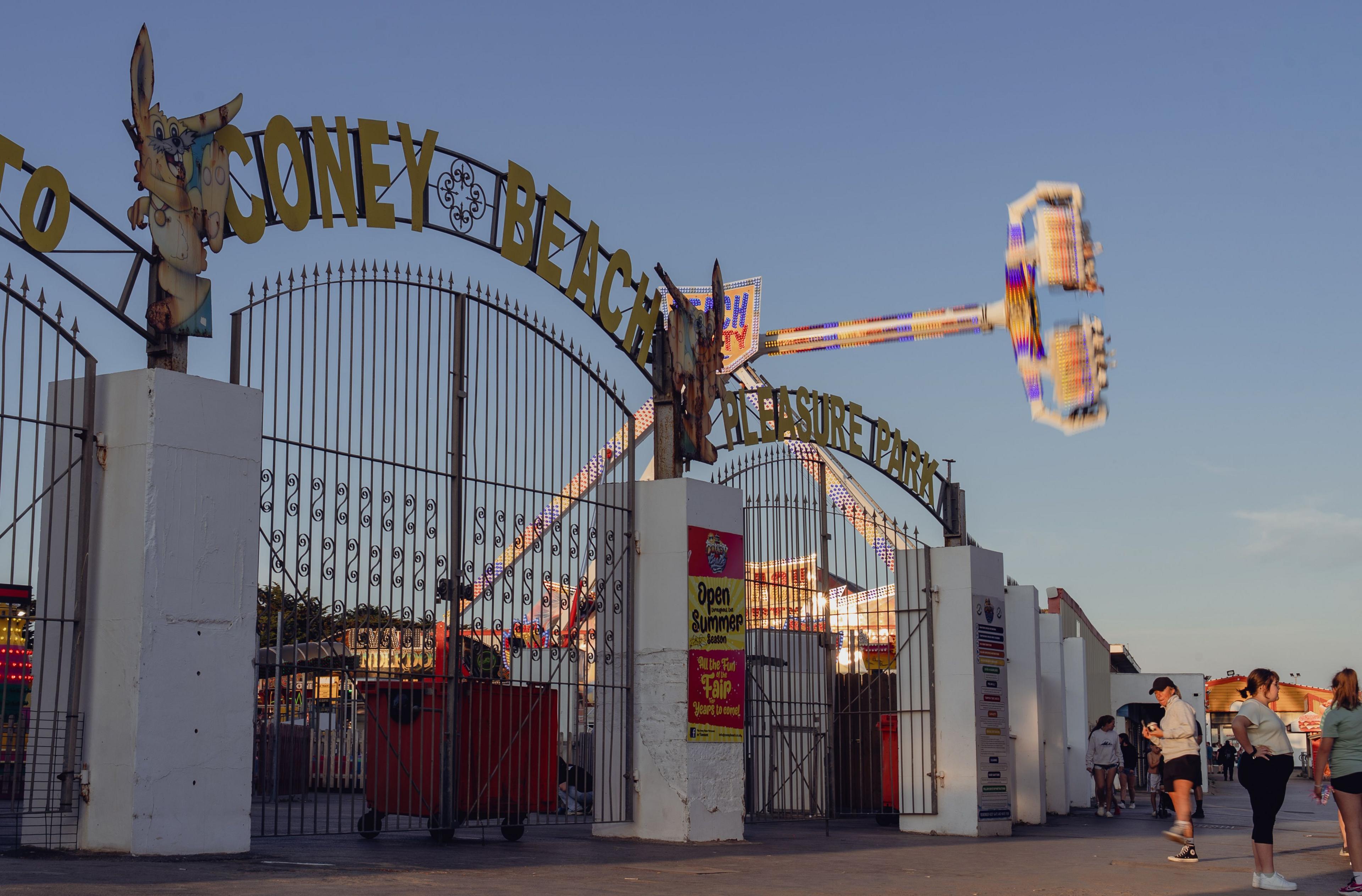 The gates to Coney Beach Amusement Park in the late afternoon sun. People are socialising in front of the entrance to a fairground with a revolving gondola ride spinning against a clear blue sky.