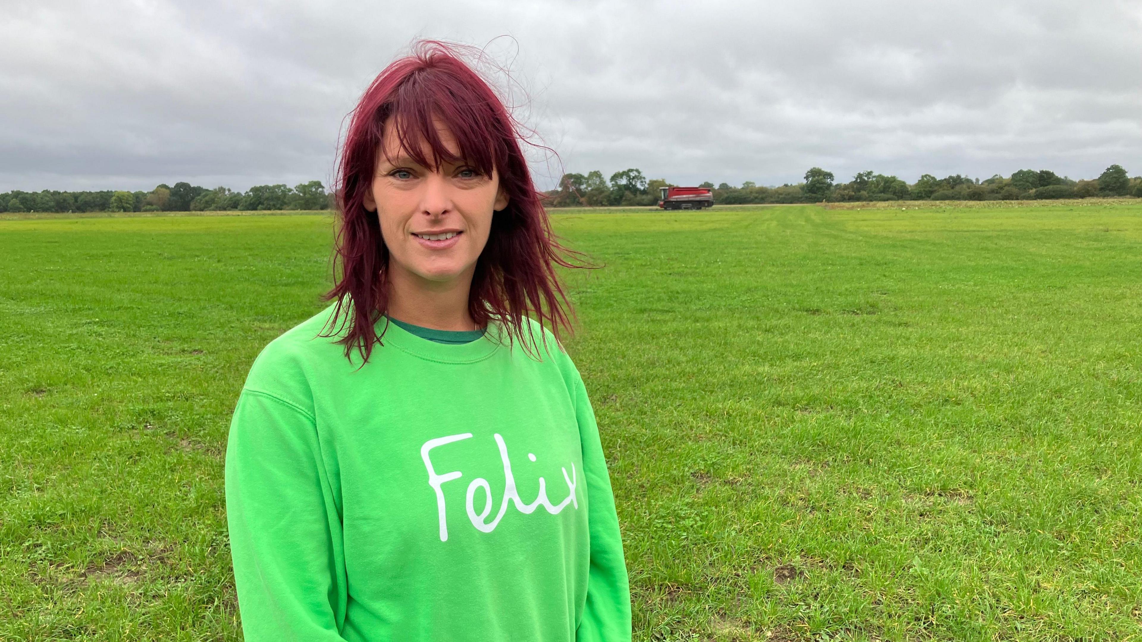 A woman with dyed red/purple hair and a green jumper stands in a field