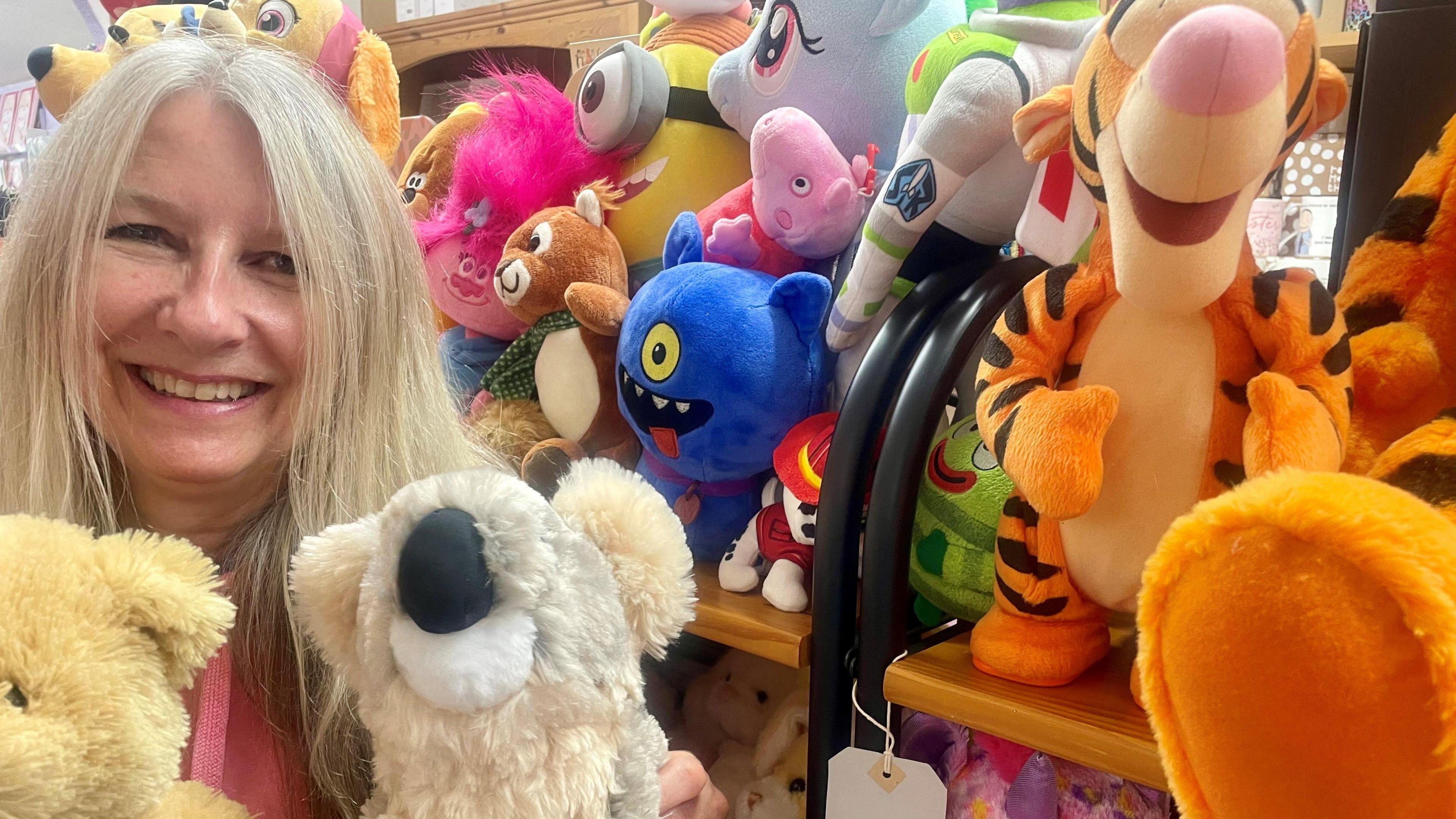 A smiling woman with blonde hair poses with two teddies in front of a shelf of soft toys, including a couple of Tiggers, Monsters Inc, Minions, Buzz Lightyear and Peppa Pig.