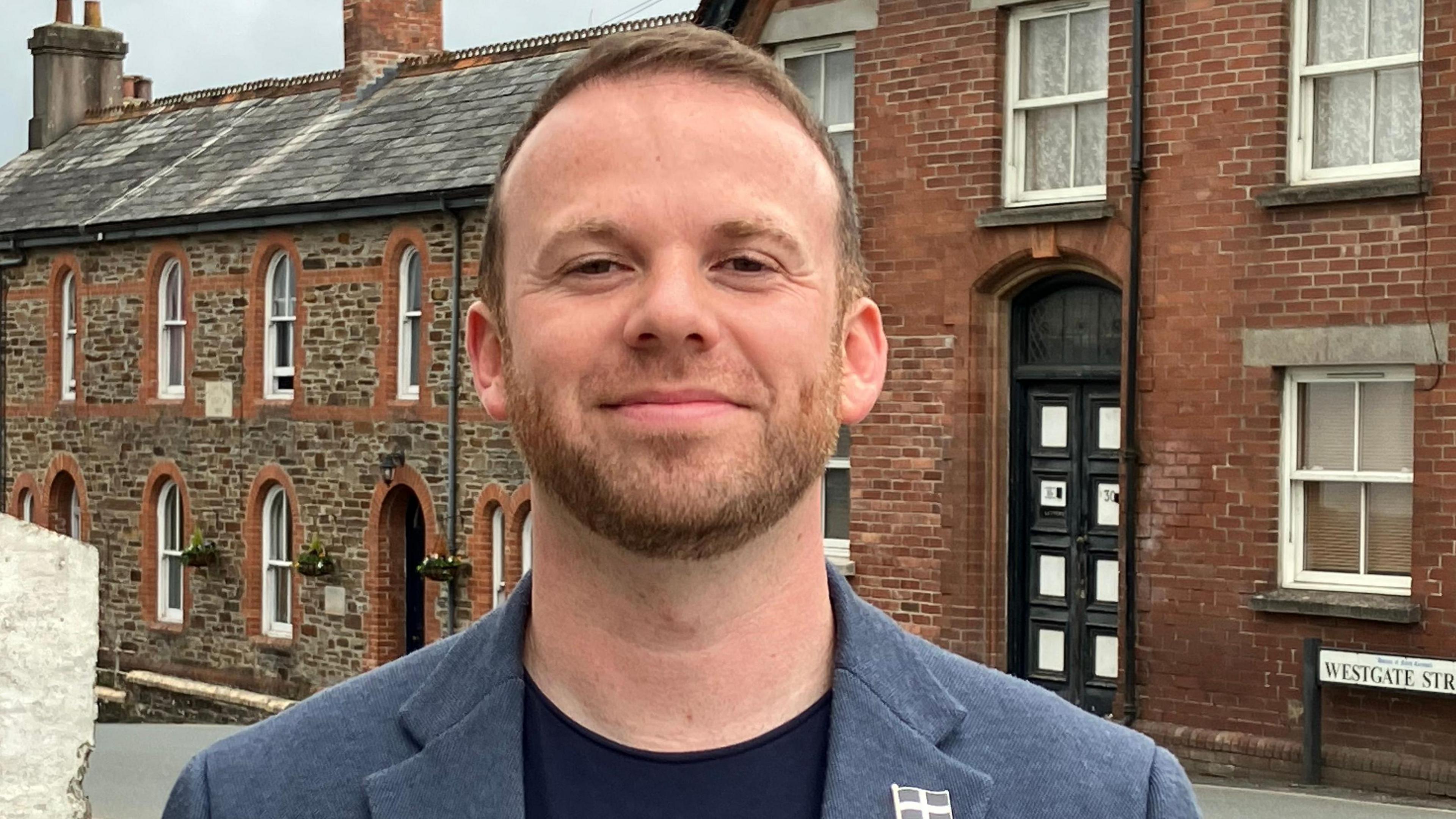 Man in dark blue t-shirt and light blue jacket with a ginger beard standing in front of a row of houses