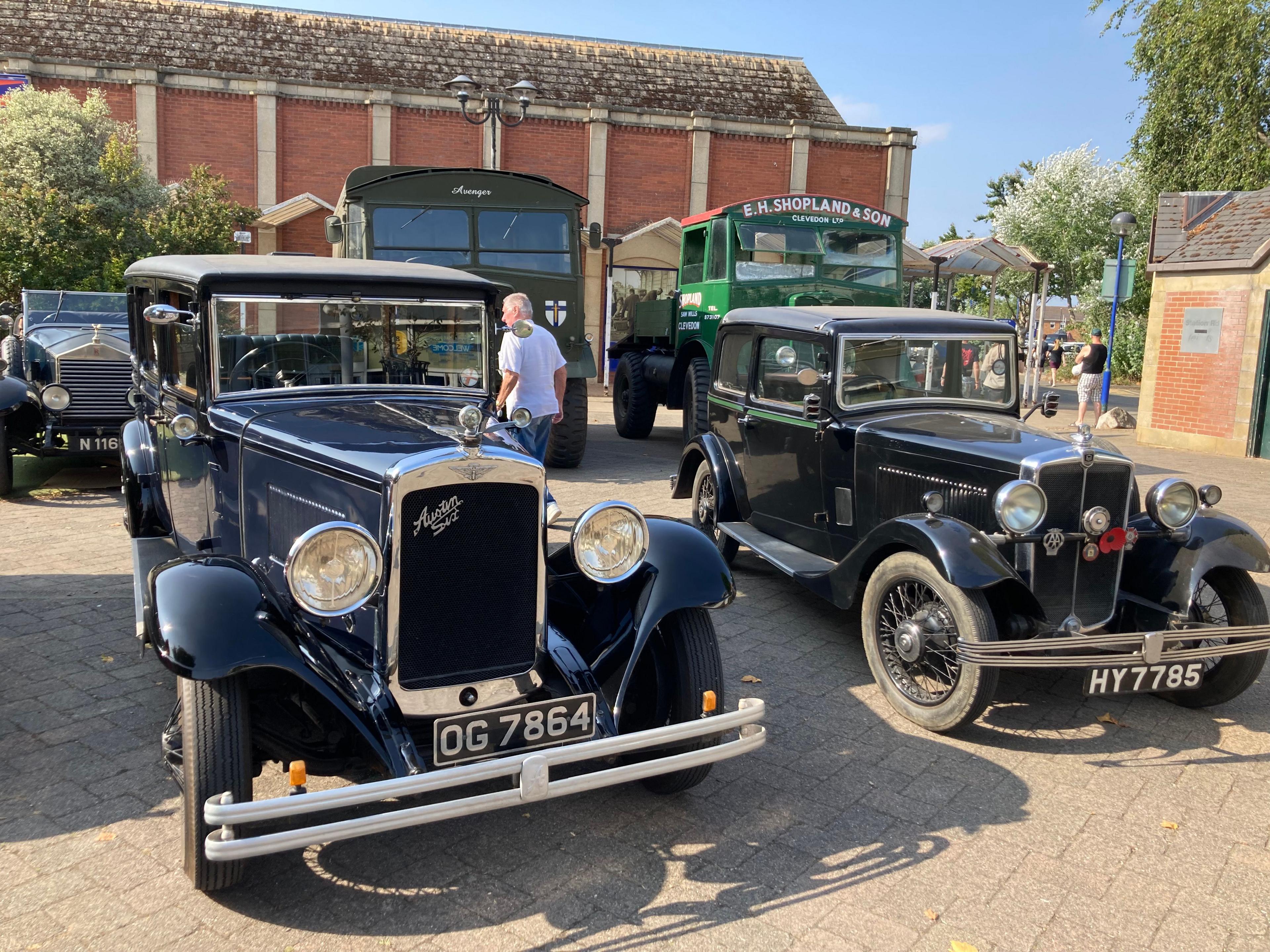 Two antique cars parked in the town centre, both of them black. They are in front of two larger vintage lorries. 