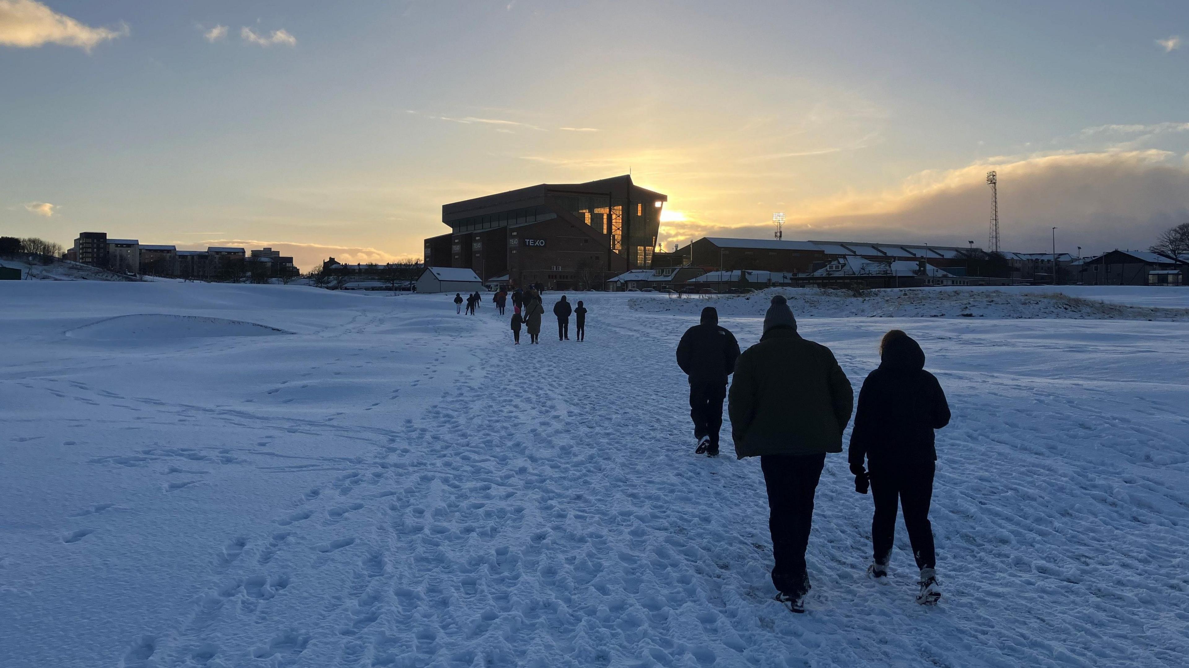 People walking away from camera towards Pittodrie Park, there is a good covering of snow and the sun is setting behind the stadium