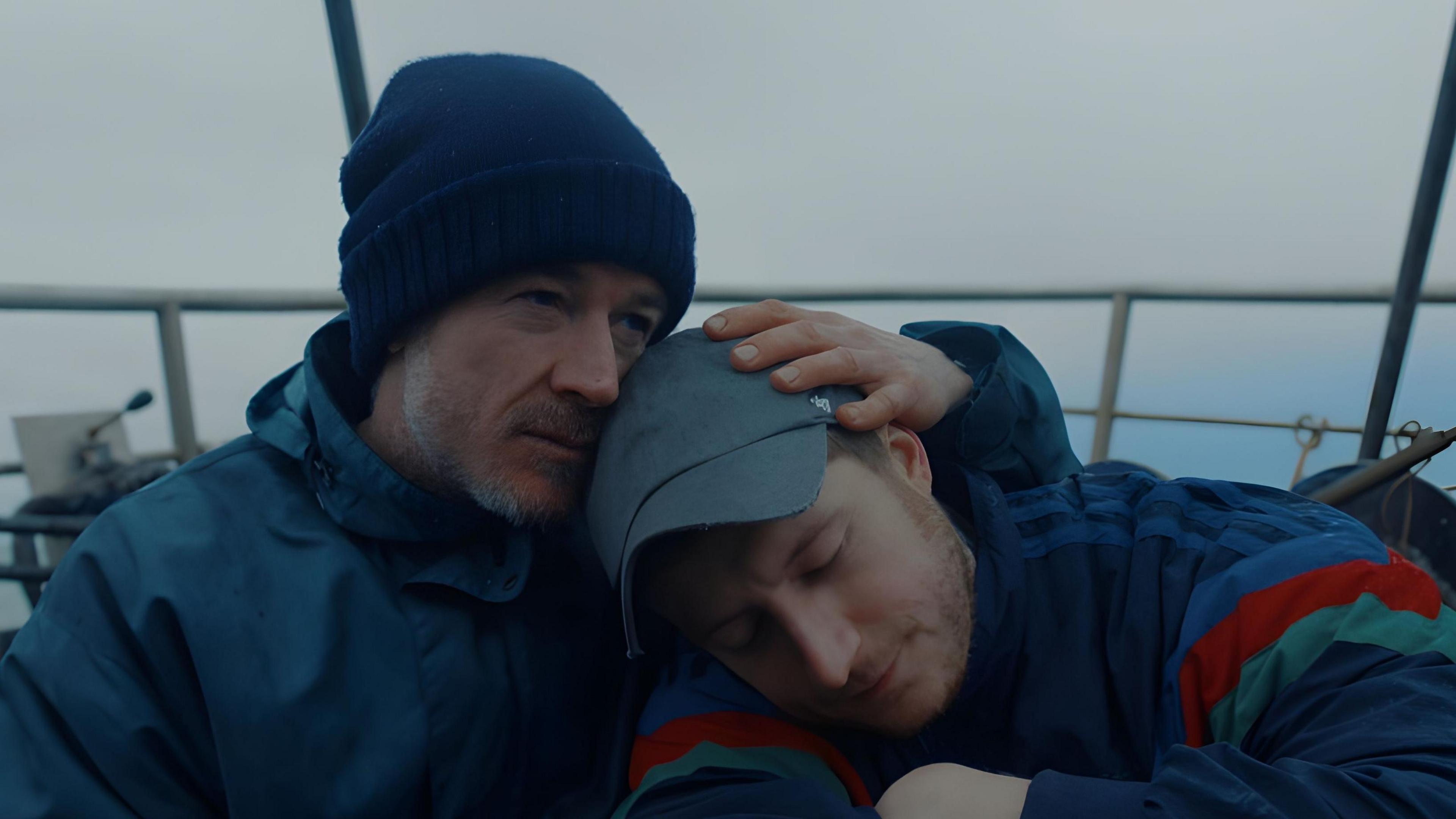 Daniel wears a blue cap and blue jacket with blue, red and green stripes on his arms. He rests his head on Jack's shoulder. Jack rests his hand on Daniel's head and his face in his head. Jack wears a blue wool hat and blue waterproof coat. 