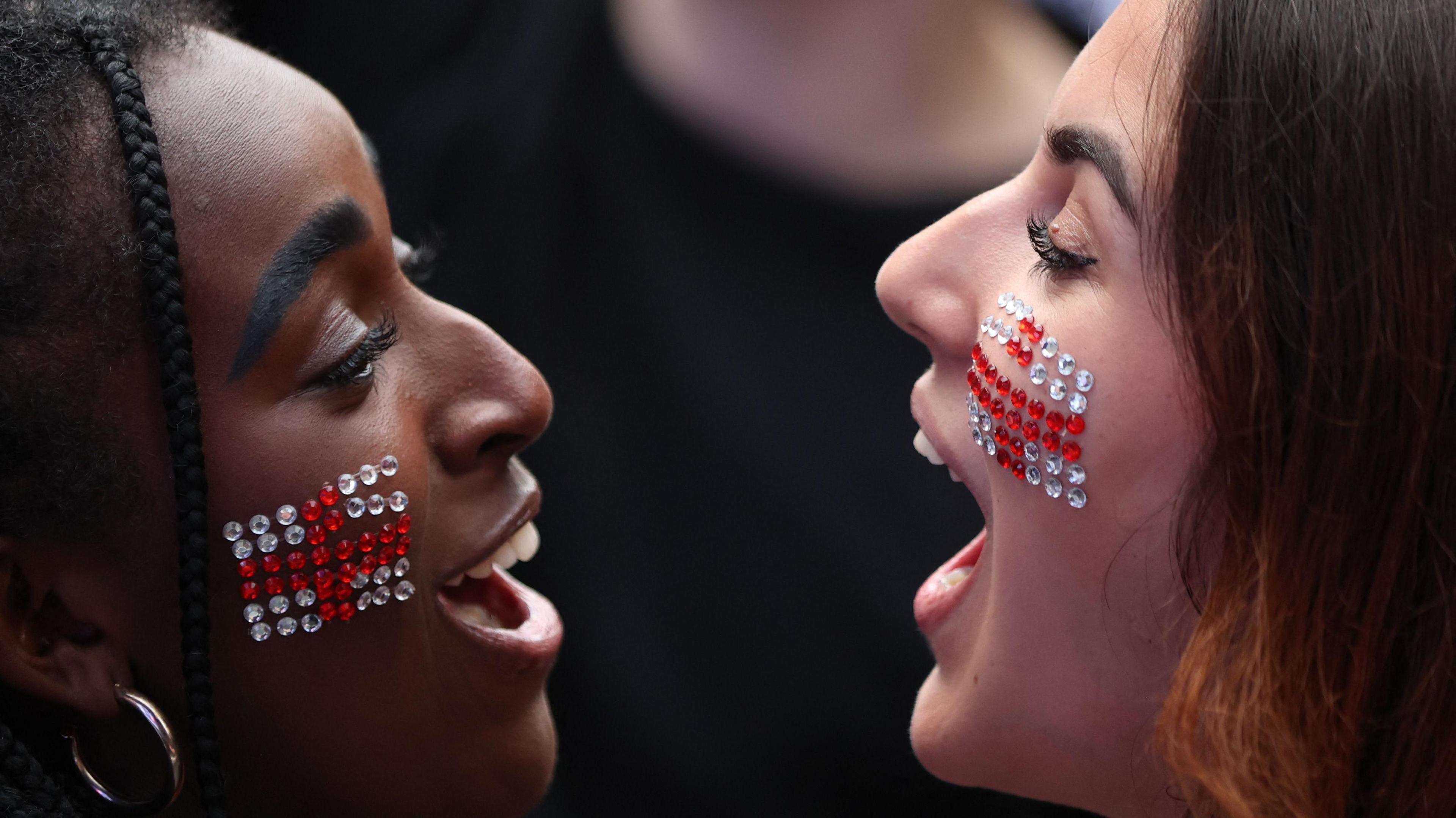 Two girls with the England flag in sequins on their cheeks look at each other